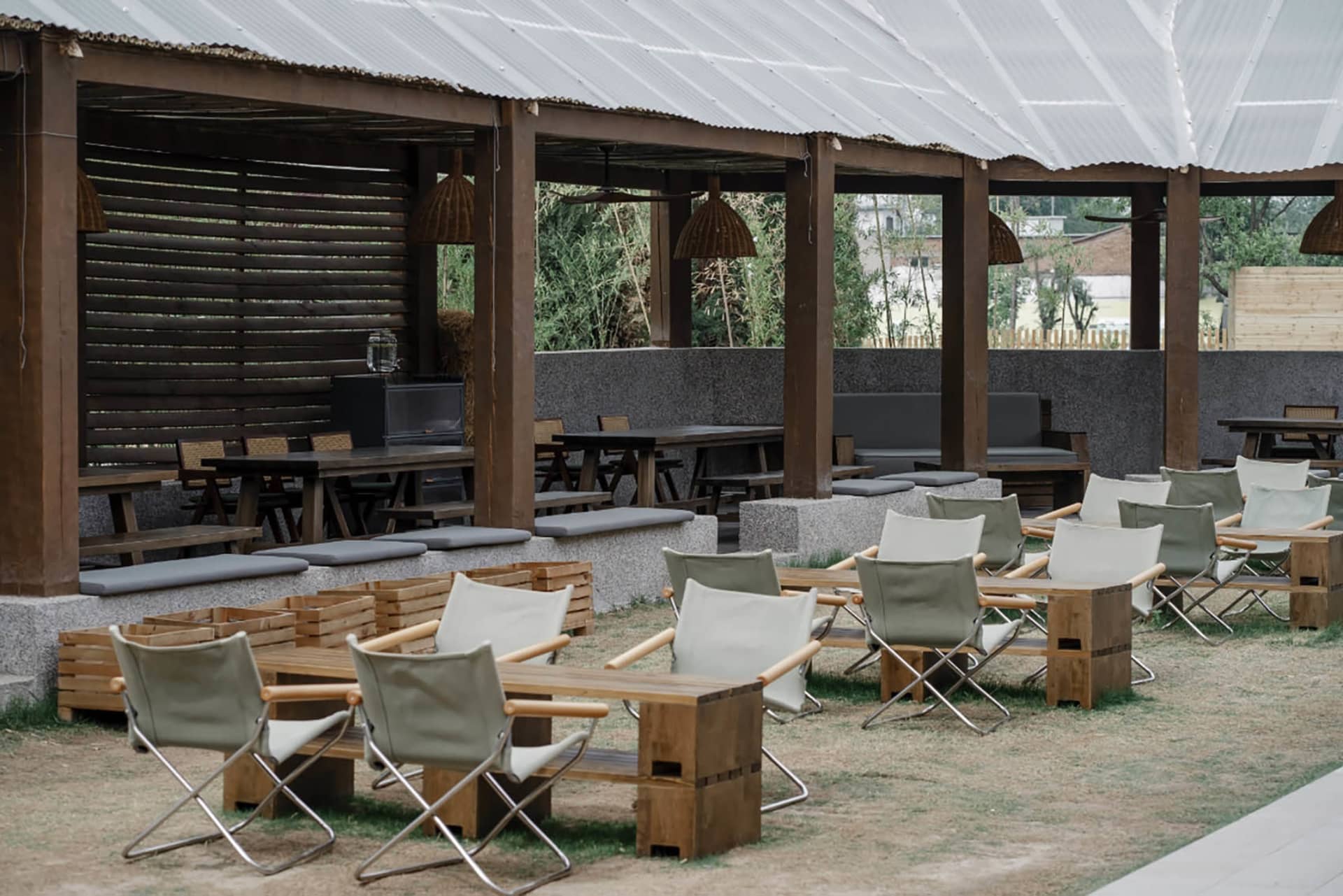 A restaurant patio area with wooden tables and chairs under a large umbrella, set on a dirt ground with a garden-like atmosphere