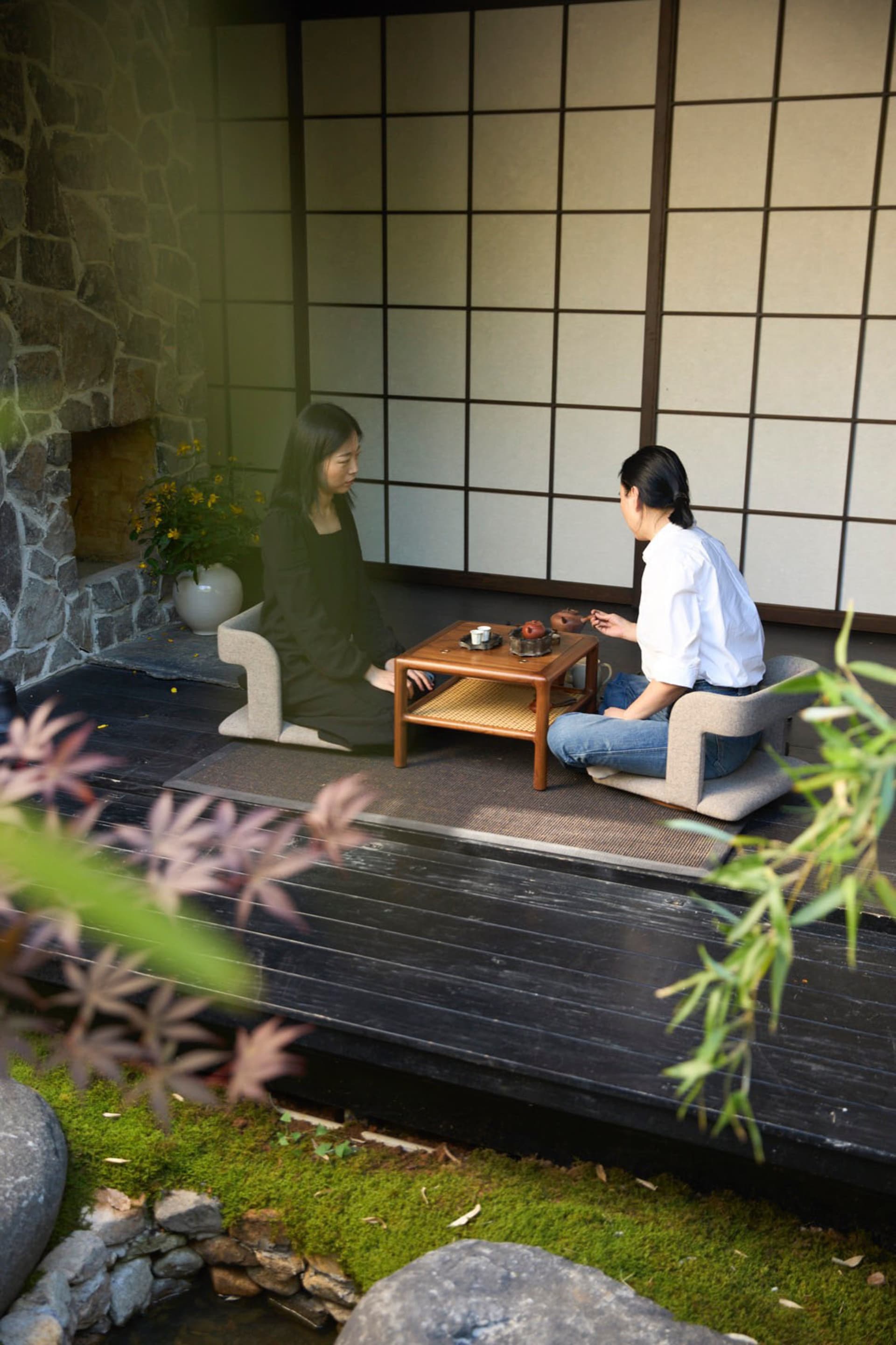 兩位女性坐在亞洲風格客廳中，木質桌子上擺放裝飾花瓶與盆栽，營造舒適氛圍