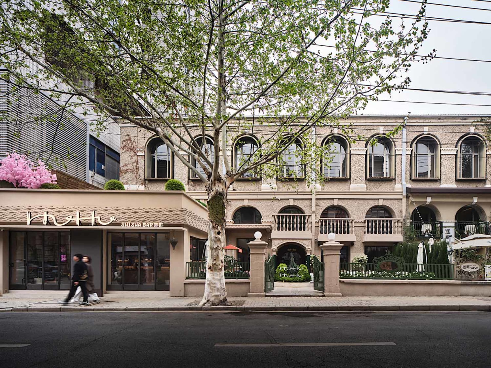 A rendered scene of a building with a brick facade, surrounded by potted plants, umbrellas, and a tree, set in a public space with a traditional atmosphere