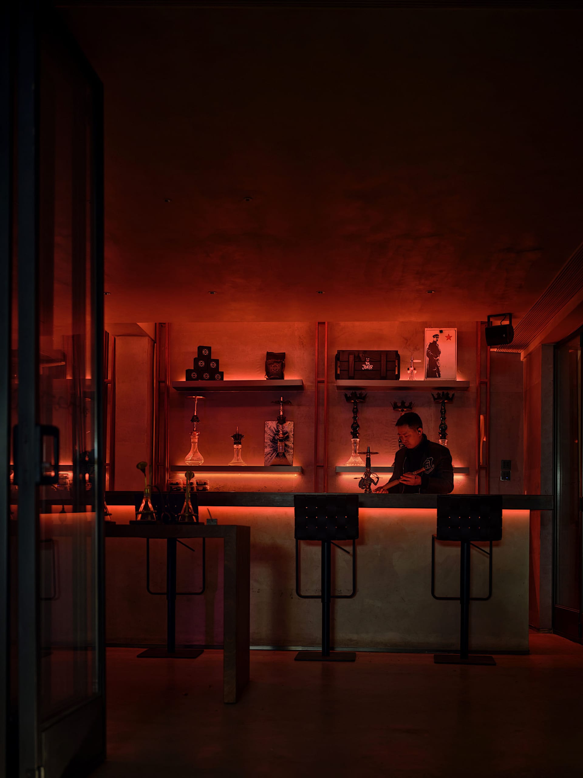 Dimly lit bar area with a stone-like wall, a counter, and stools, featuring bottles and a modern ambiance