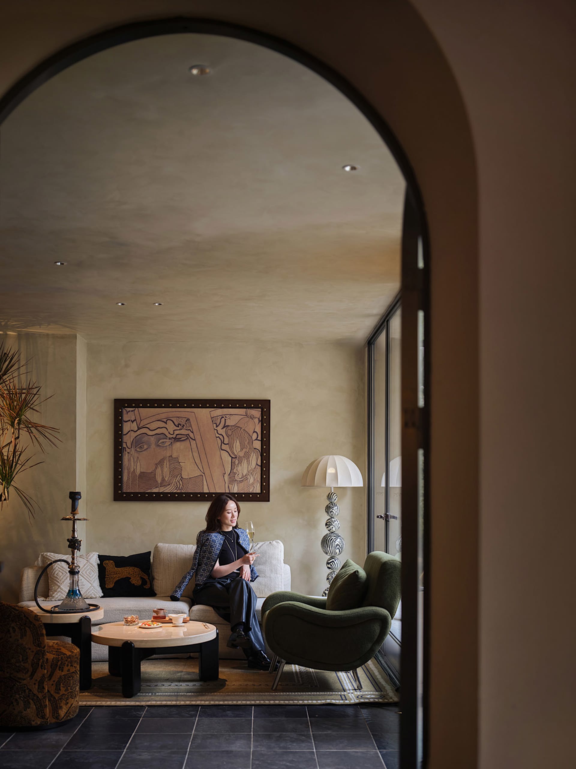 A woman sits in a cozy living room with a dining table, couches, a potted plant, and a vase, surrounded by warm and inviting decor