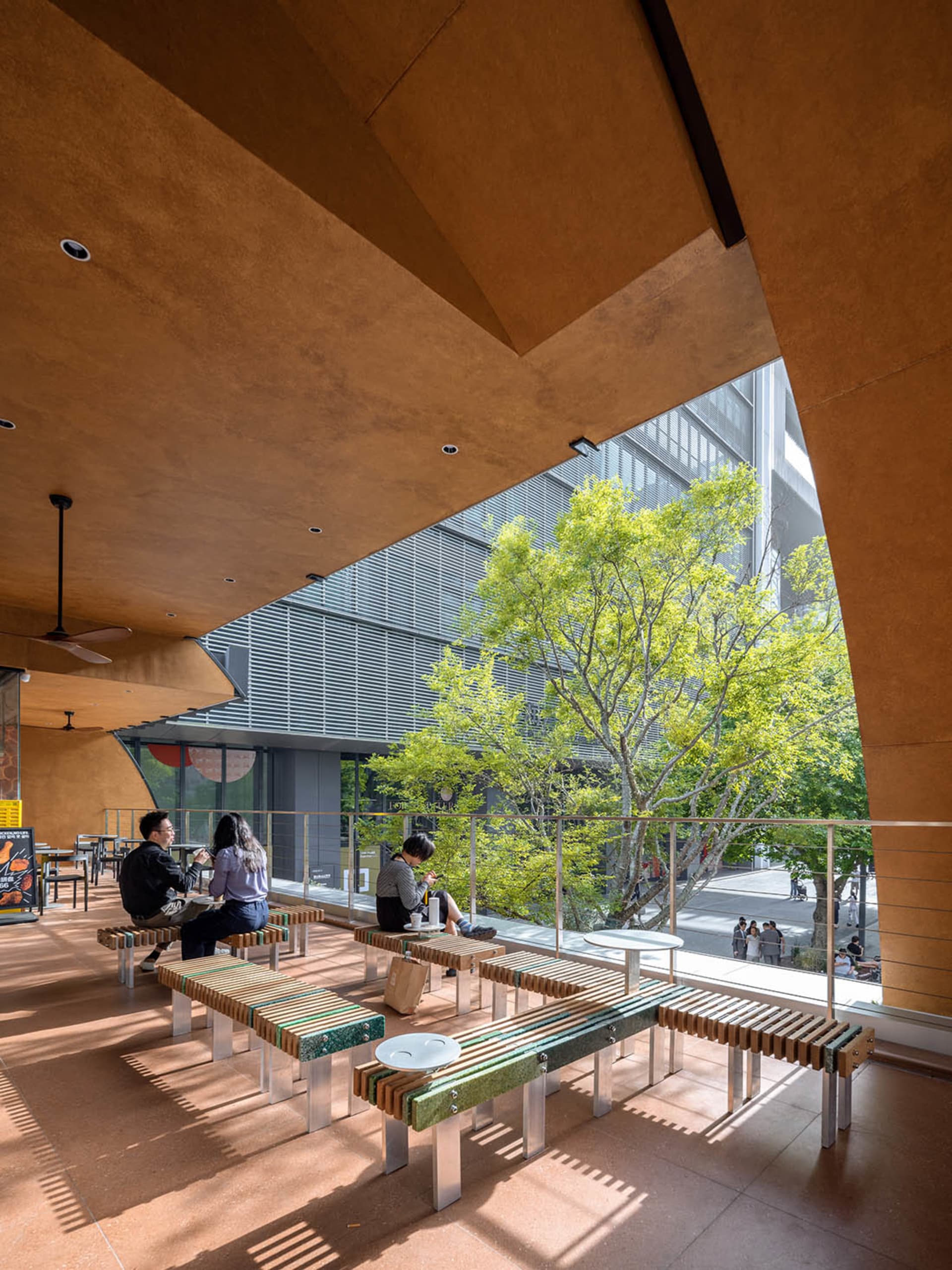 Dining area with wooden benches and tables, natural light from large windows, and several people enjoying meals in a warm and welcoming atmosphere