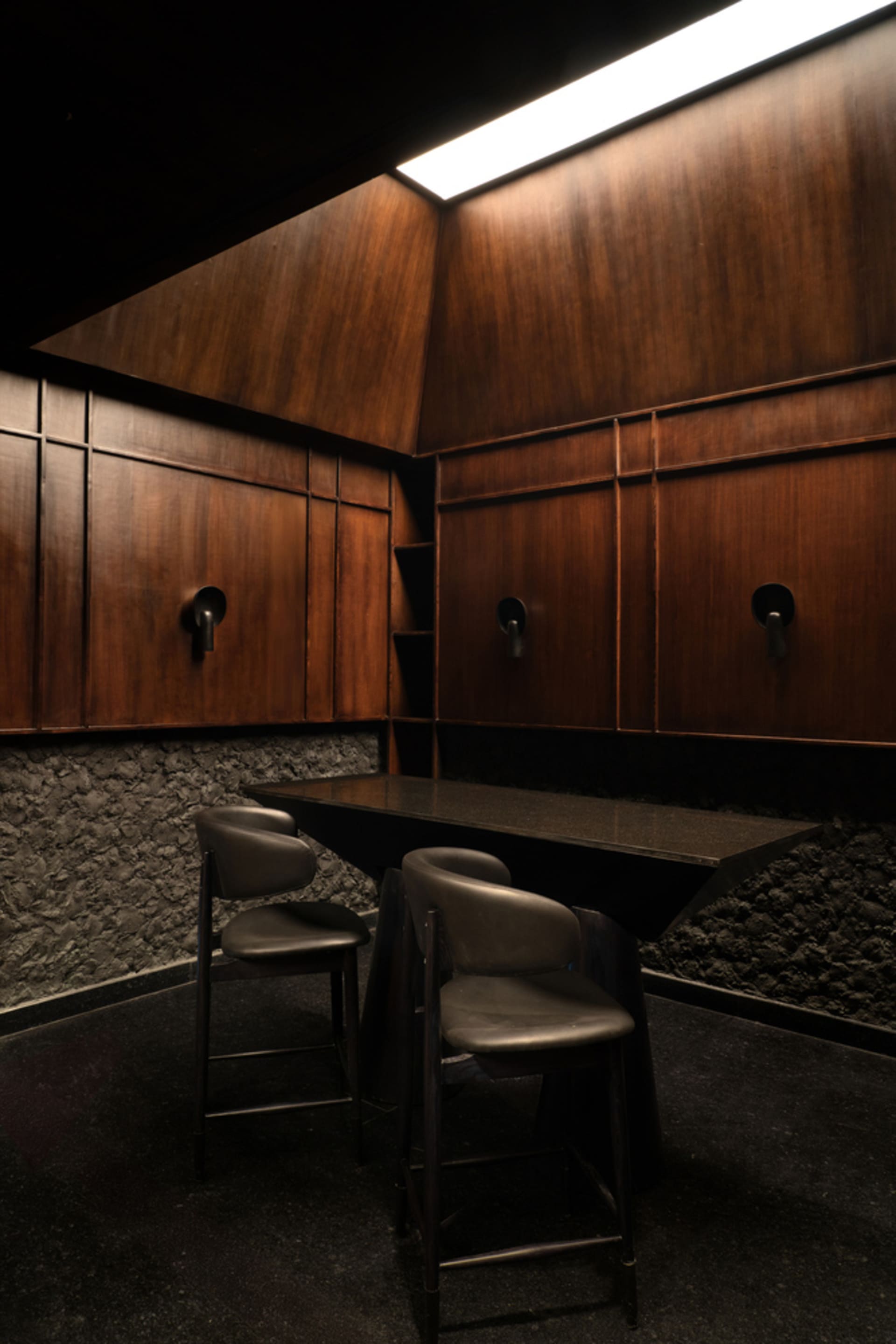 A modern restaurant or bar area with wooden walls and floors, featuring two black leather chairs and a marble dining table, well-lit with natural or artificial light