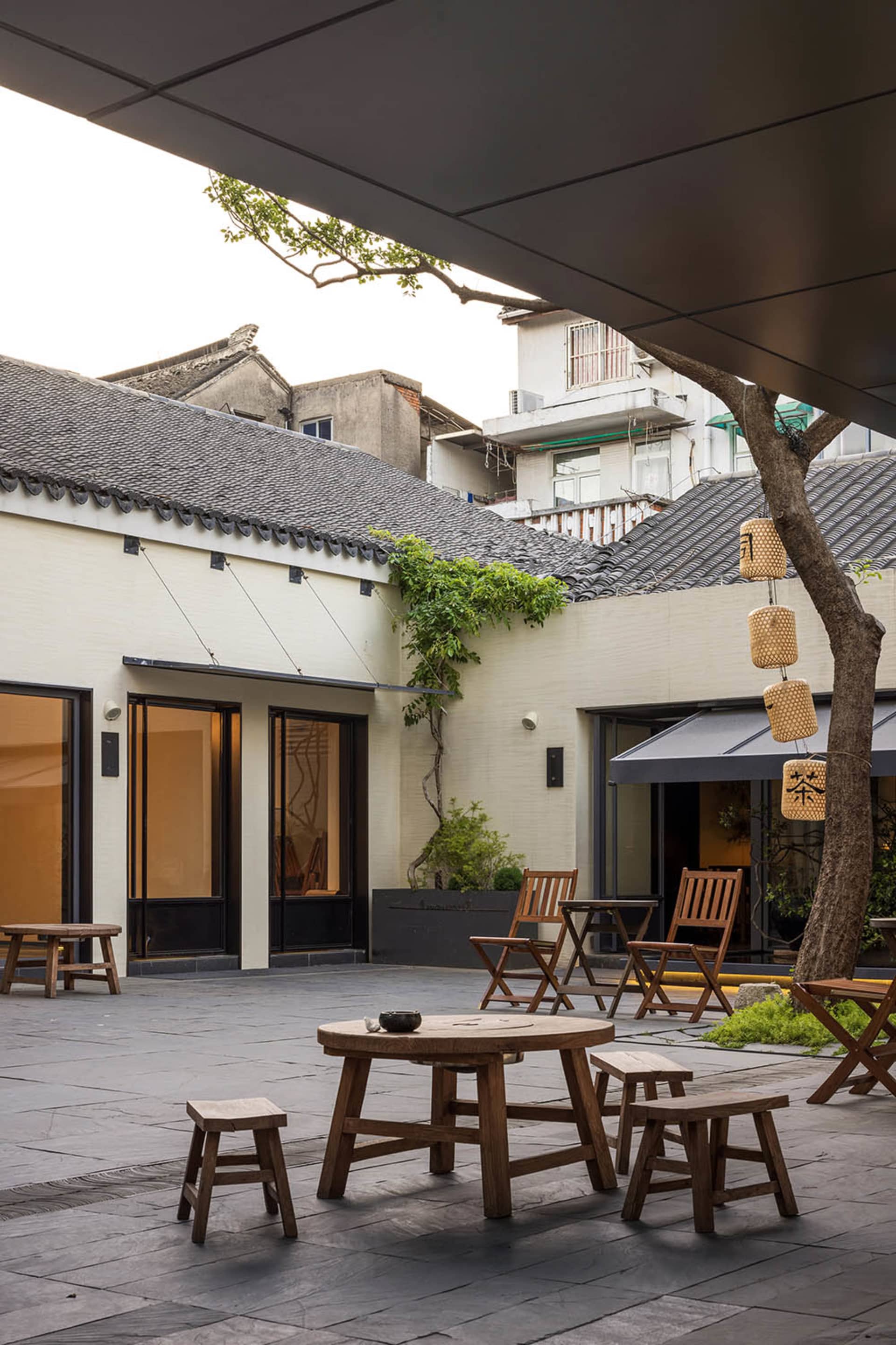 A welcoming courtyard dining area with wooden chairs, a dining table, and potted plants, set within a larger building complex