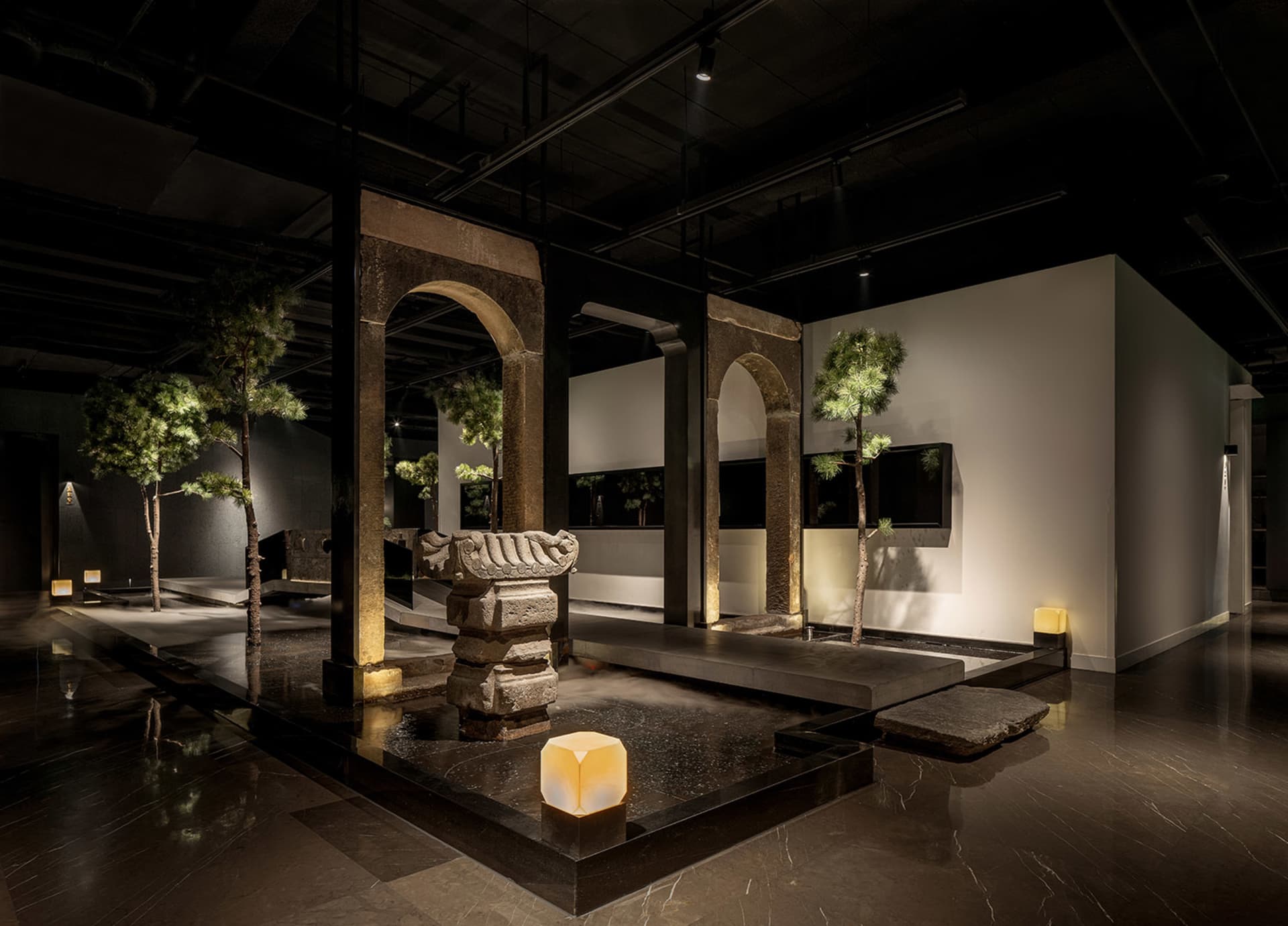 A calm and inviting art gallery space featuring a large stone sculpture, potted plants, and two vases, with a focus on the impressive stone centerpiece