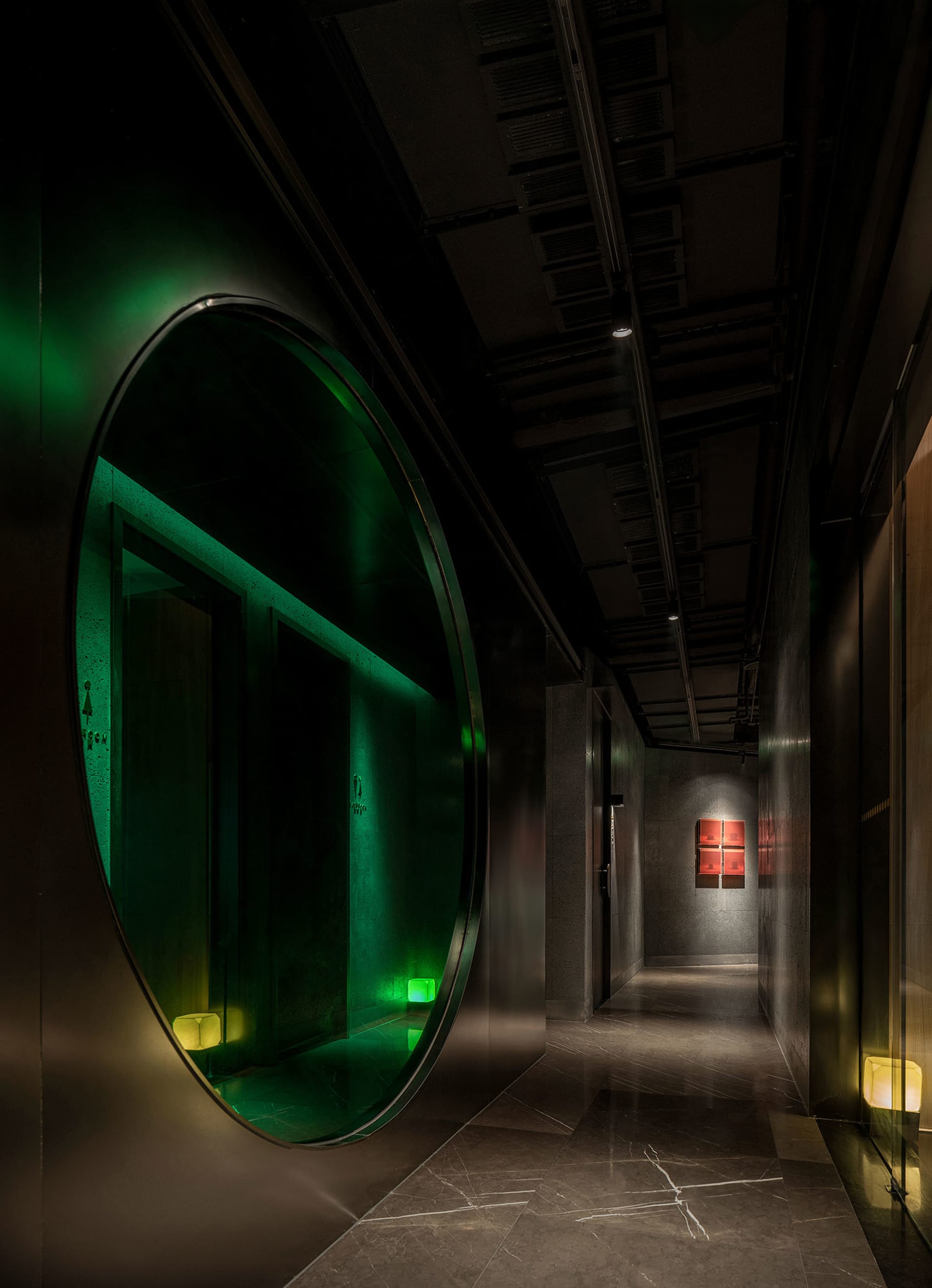 Industrial hallway with a circular mirror, concrete walls, and a painting on the wall, lit by two lights