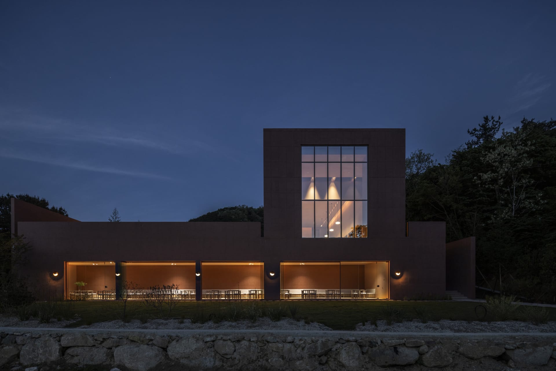 A modern building at night with a large window, stone and glass elements, and several chairs in a dining area or waiting lounge