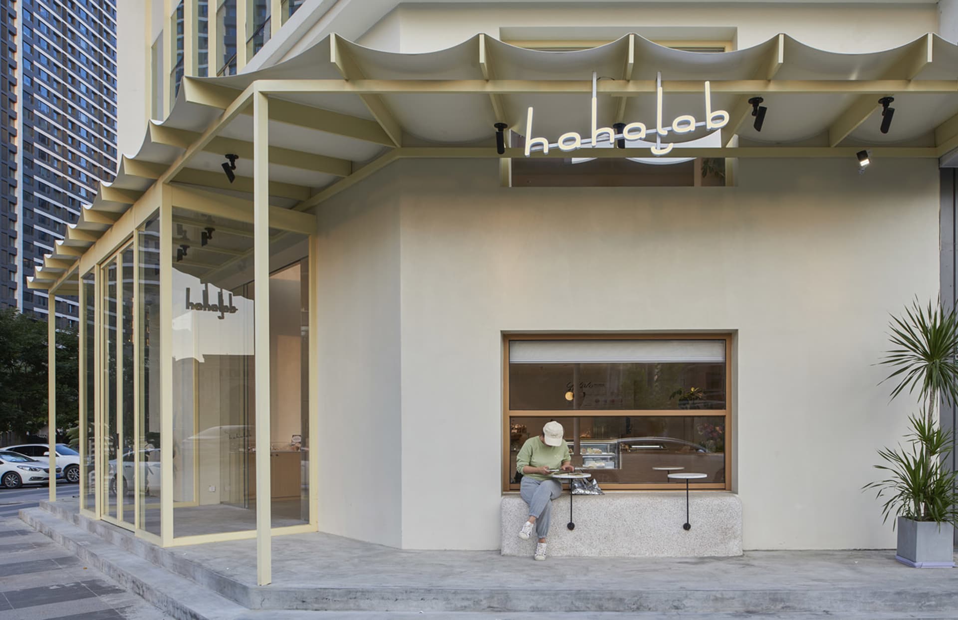 A white storefront with a yellow awning and potted plants, with a woman sitting on a bench outside and a car parked in the background