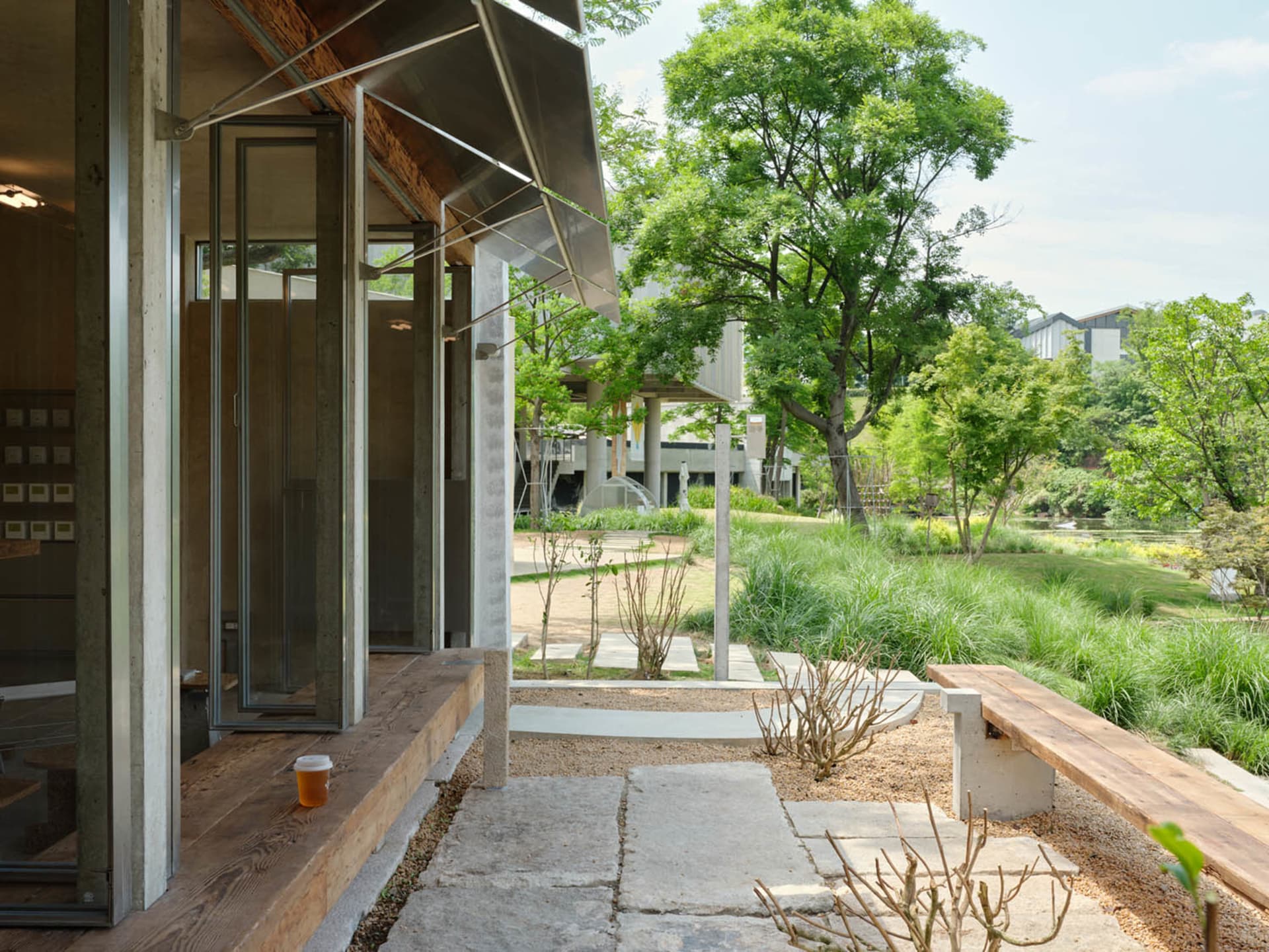 A serene outdoor space with a modern building featuring concrete walls, a metal roof, and a patio area with a bench, dining table, and potted plants