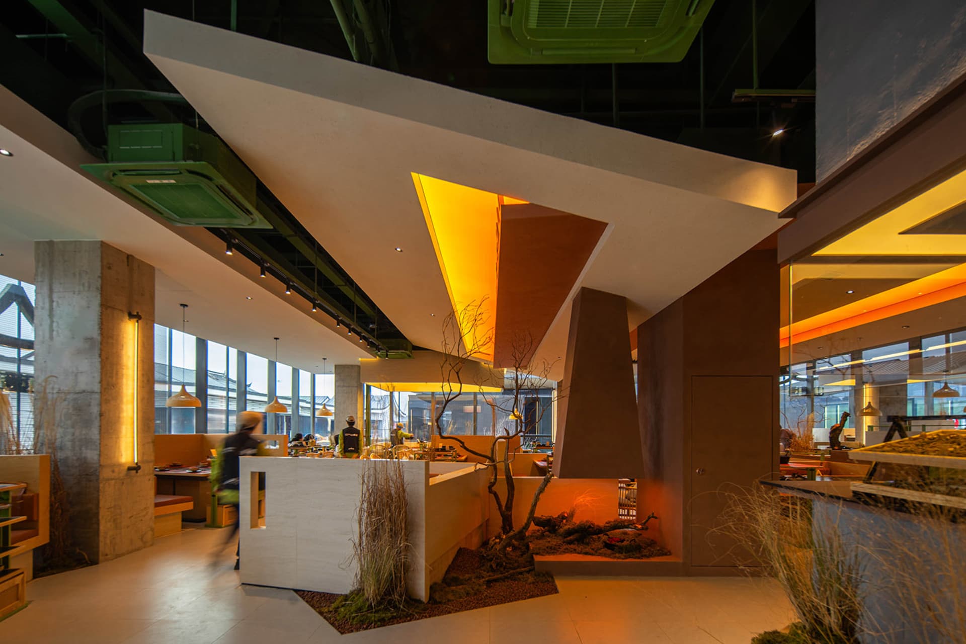 A modern restaurant interior with brown and orange tones, dining tables, chairs, potted plants, and vases, with people present