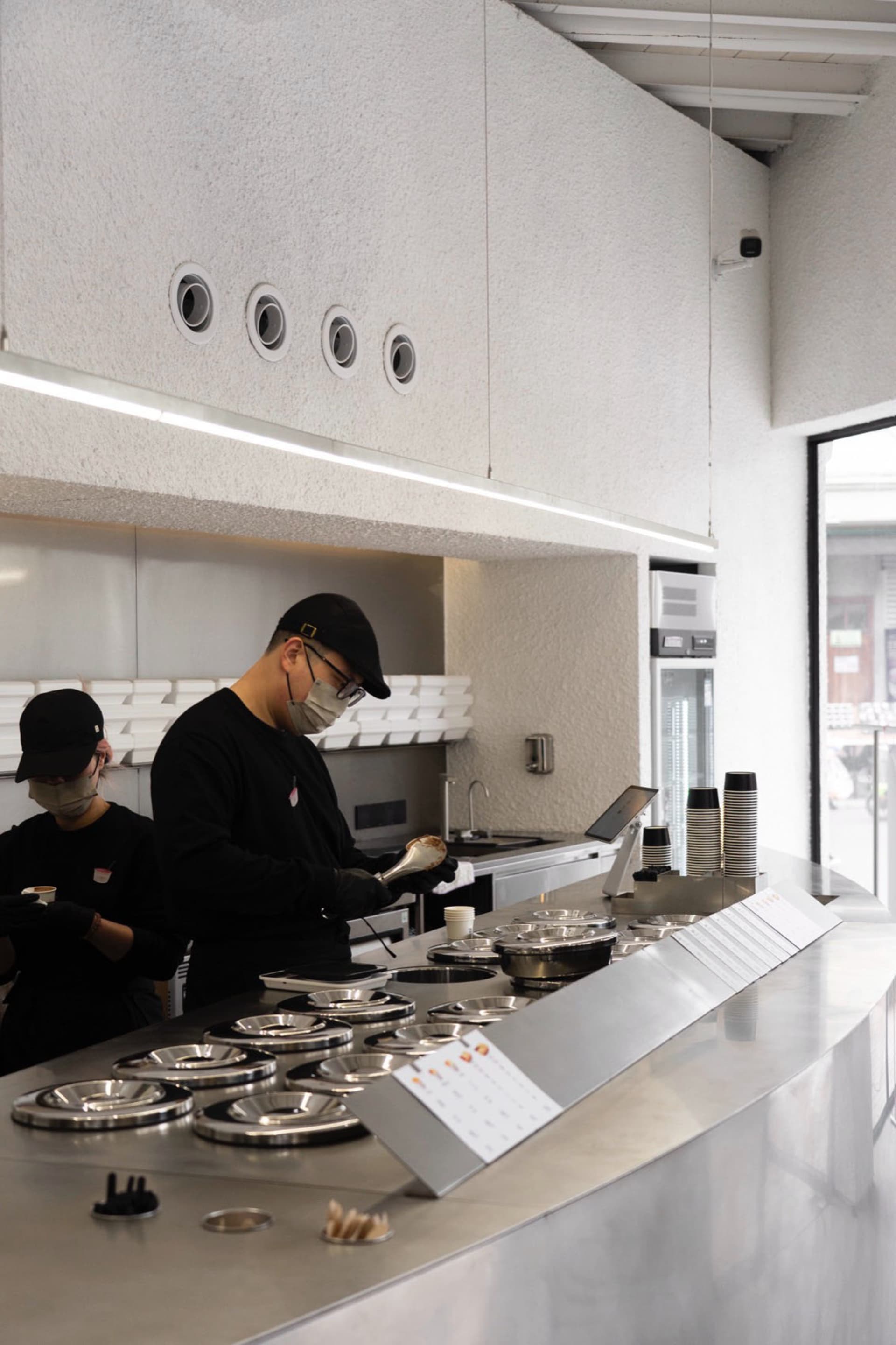 Industrial kitchen with two people working, wearing face masks, surrounded by appliances, bowls, and cups