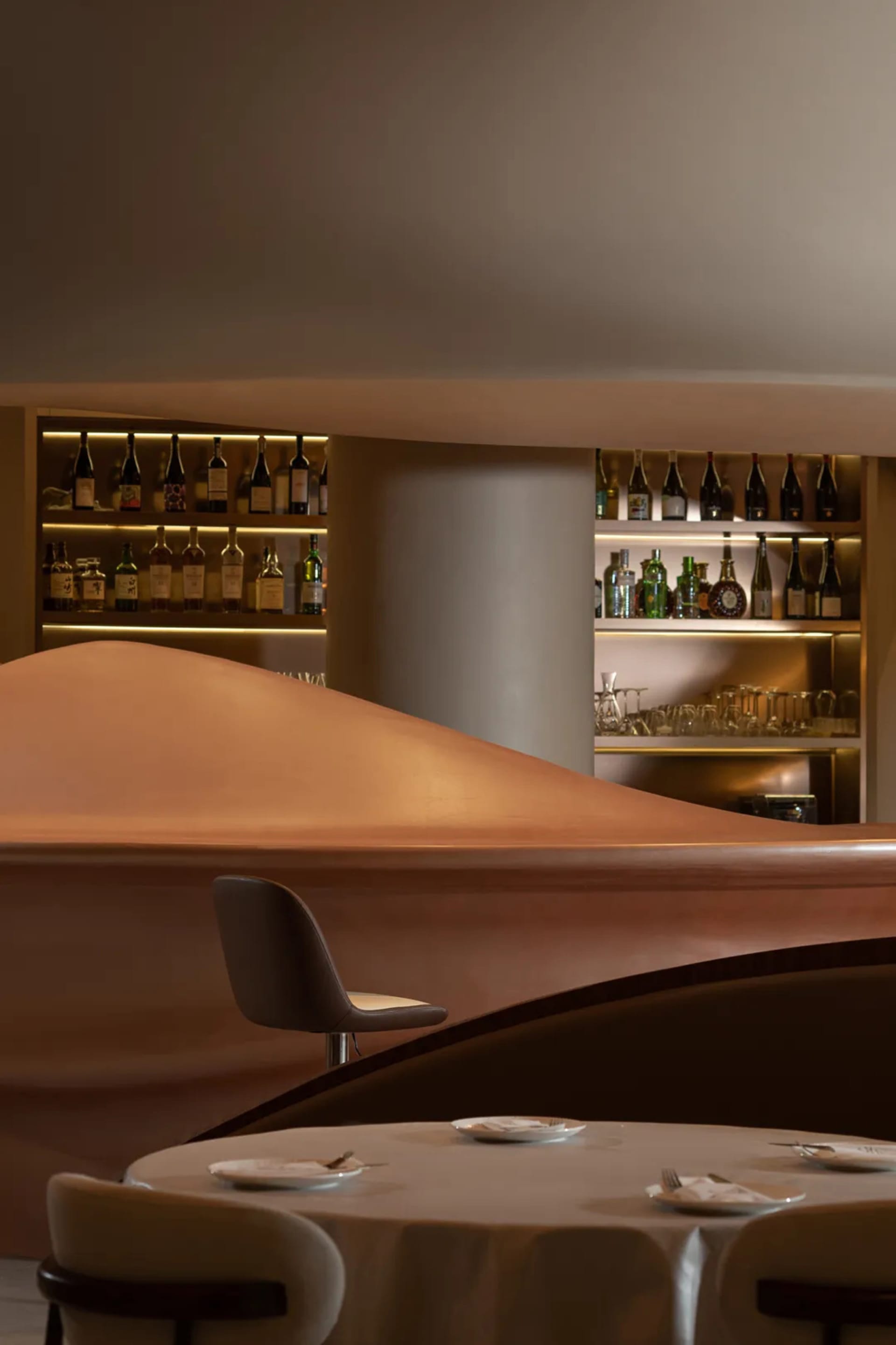A curved bar with wine glasses, cups, and bowls, surrounded by chairs for seating, in a warm and inviting atmosphere