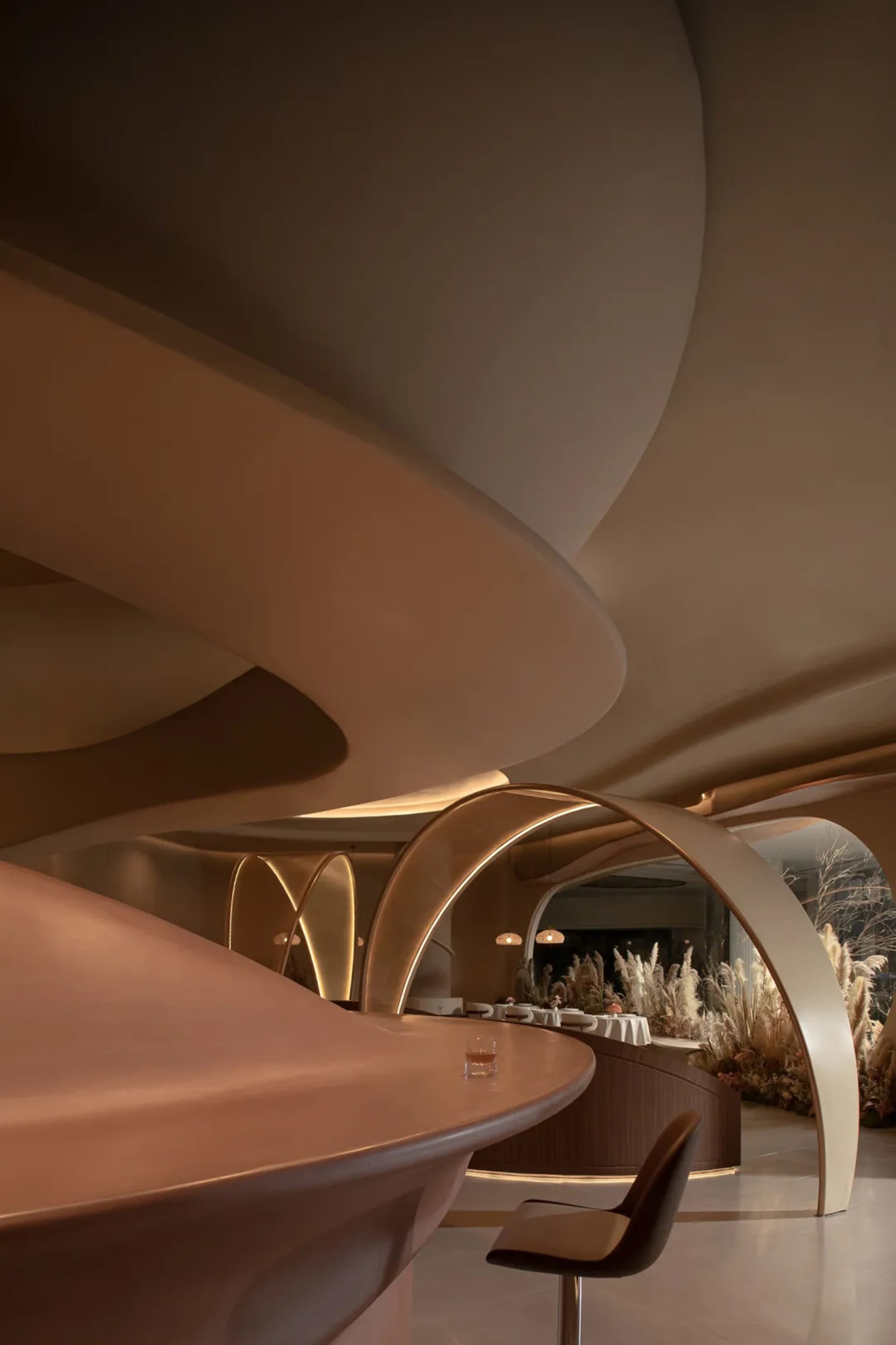 A warm and inviting restaurant interior with a curved wooden bar, chairs, and potted plants, creating a comfortable dining atmosphere