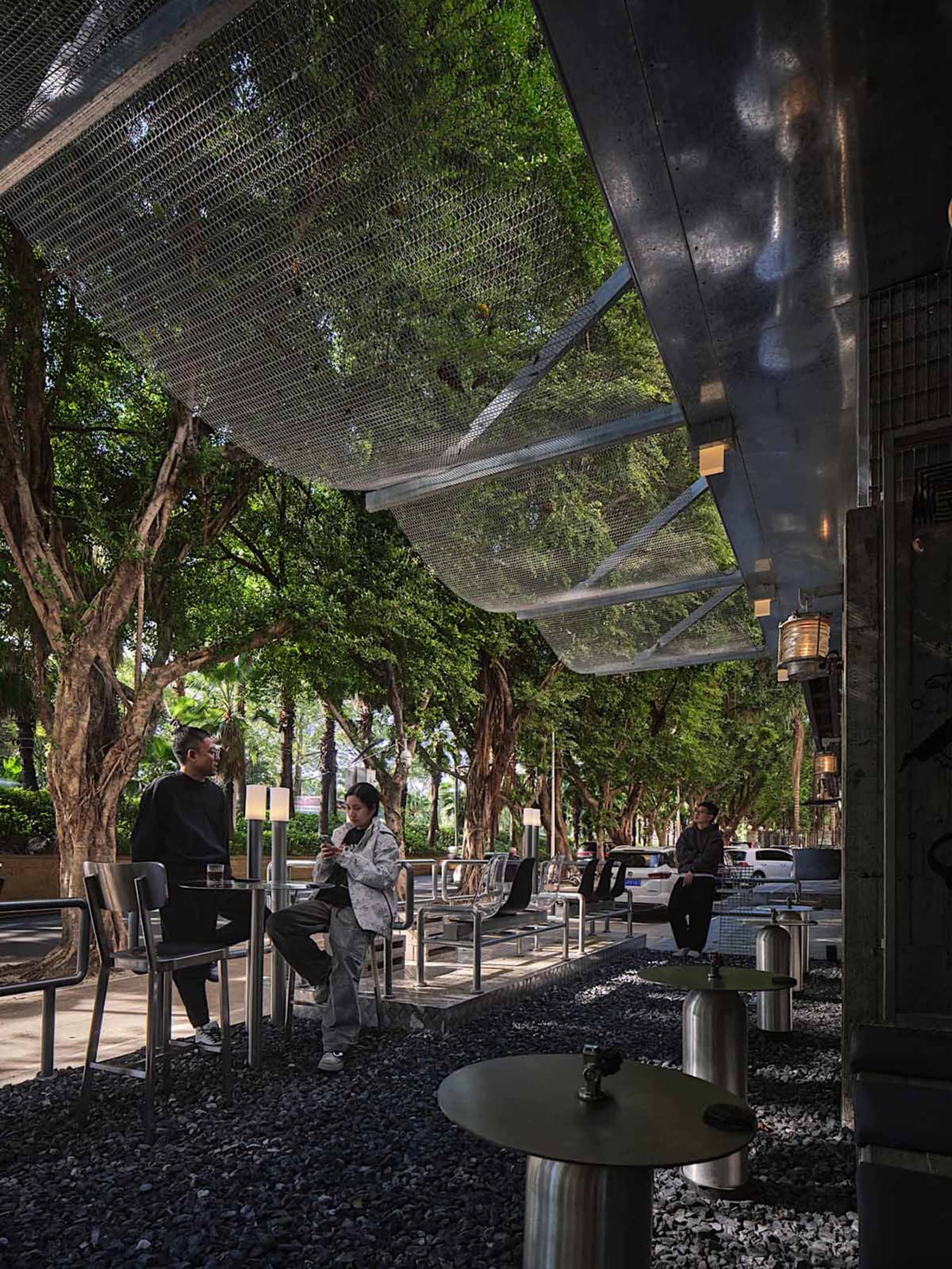 A casual outdoor seating area with metal chairs and tables under a large umbrella, surrounded by neutral tones and natural light