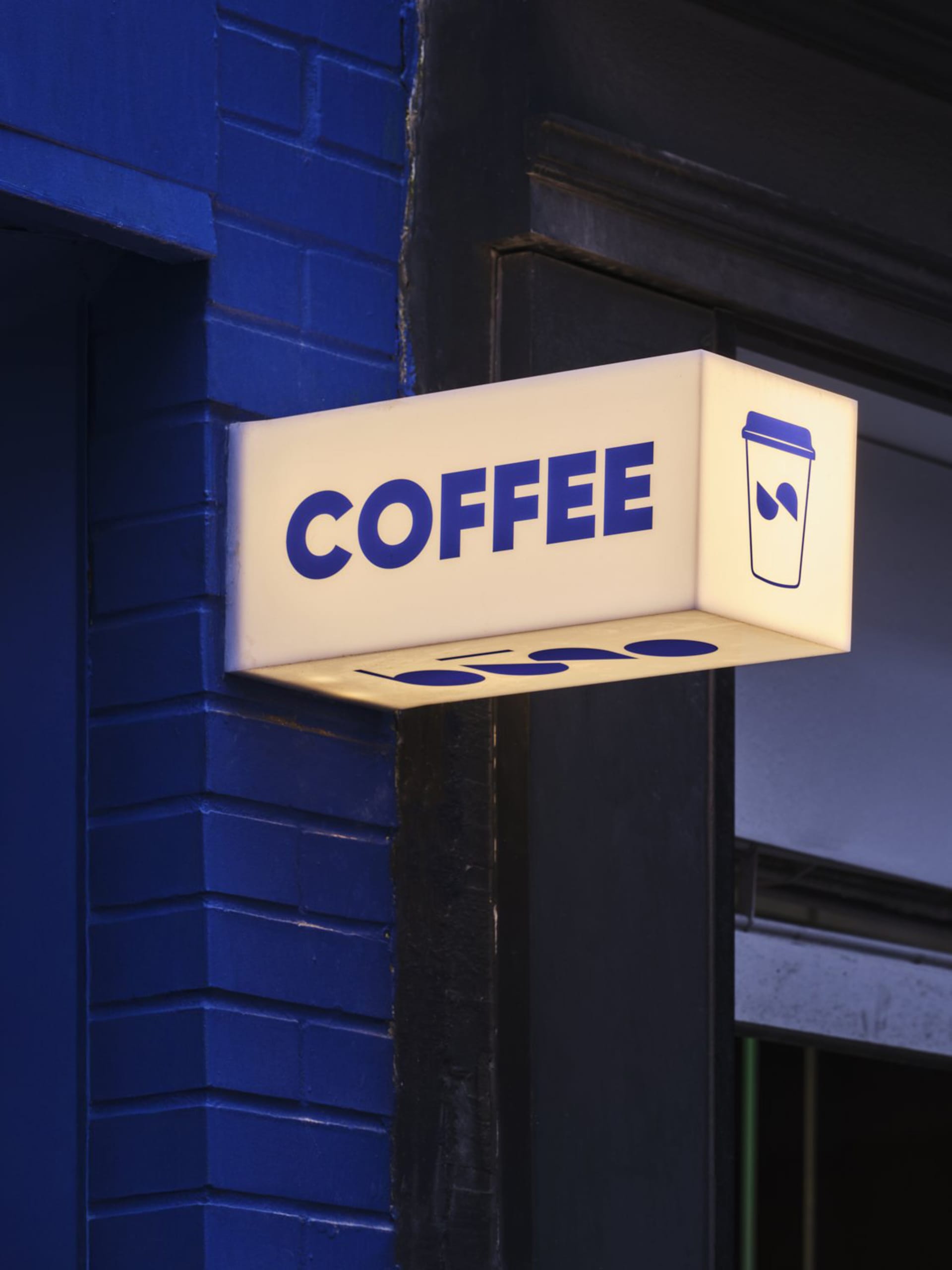 Exterior view of a coffee shop with a brightly lit sign featuring a cup and spoon, surrounded by city buildings