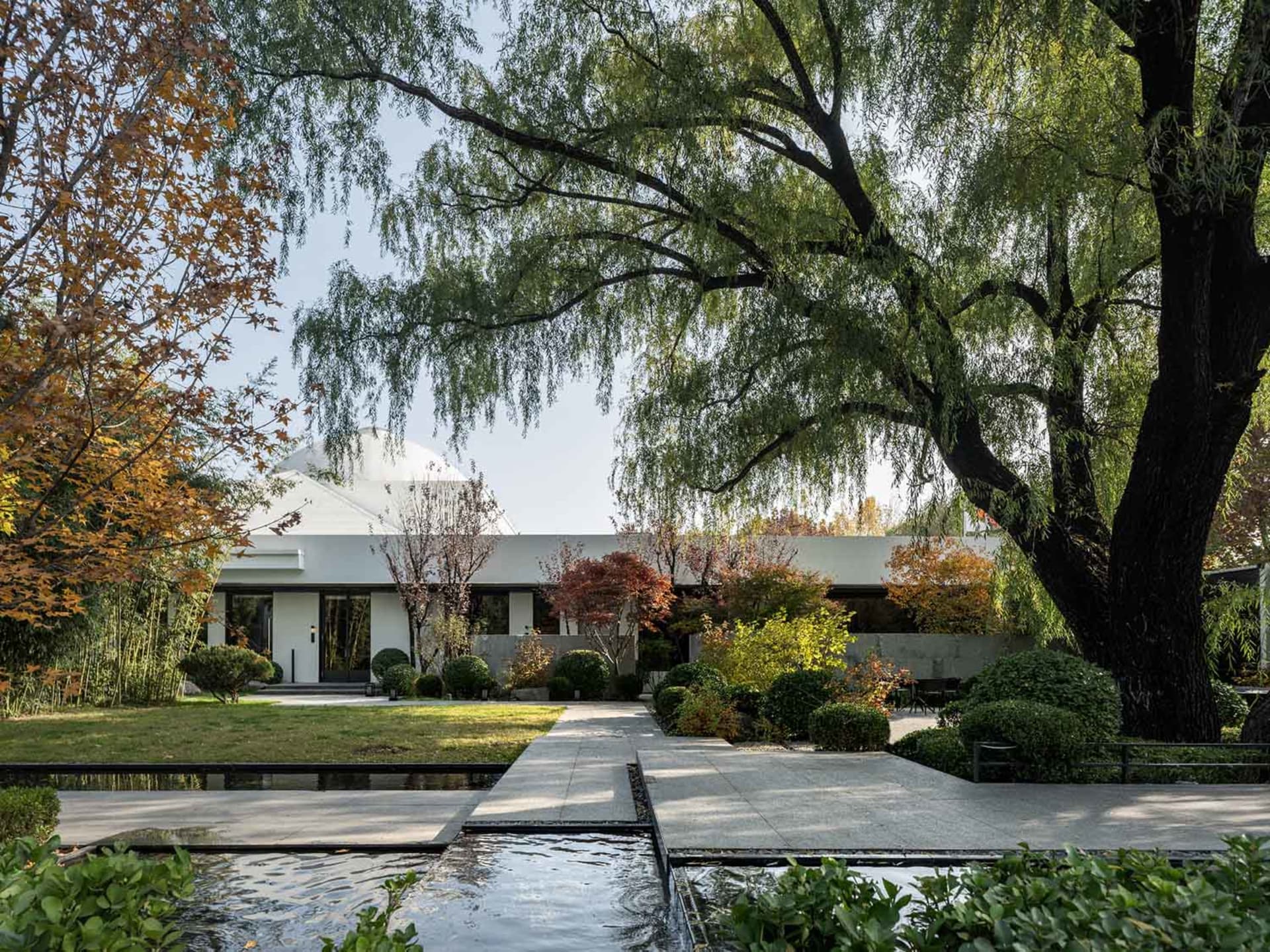 A serene architectural scene featuring a white building surrounded by trees, potted plants, and a central bench, with a mix of photo and rendered elements