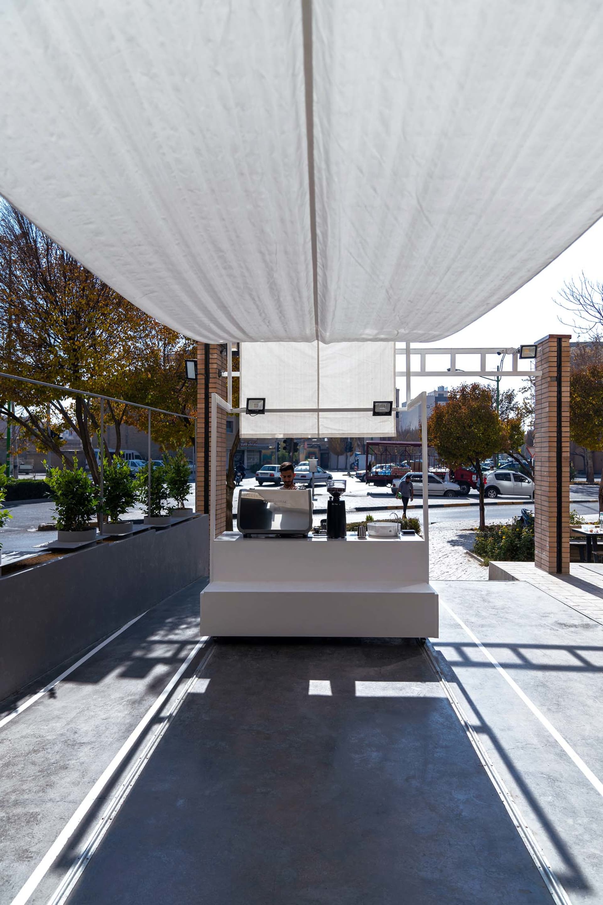 A bustling street cafe with a white counter, potted plants, and a man serving customers, surrounded by parked cars