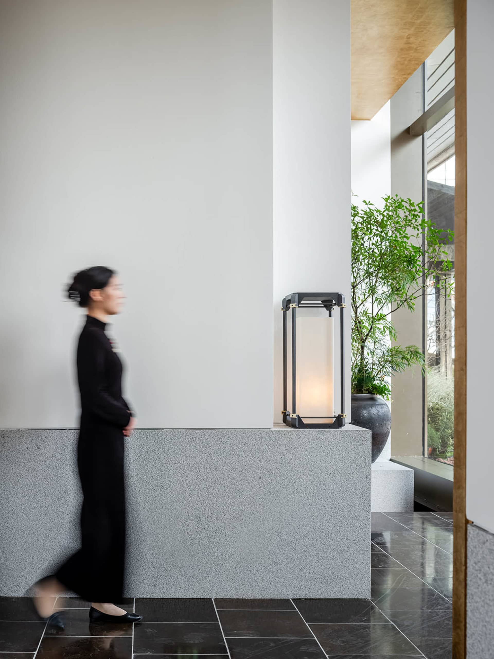 A woman walks down an office hallway with two potted plants and a suitcase on the floor