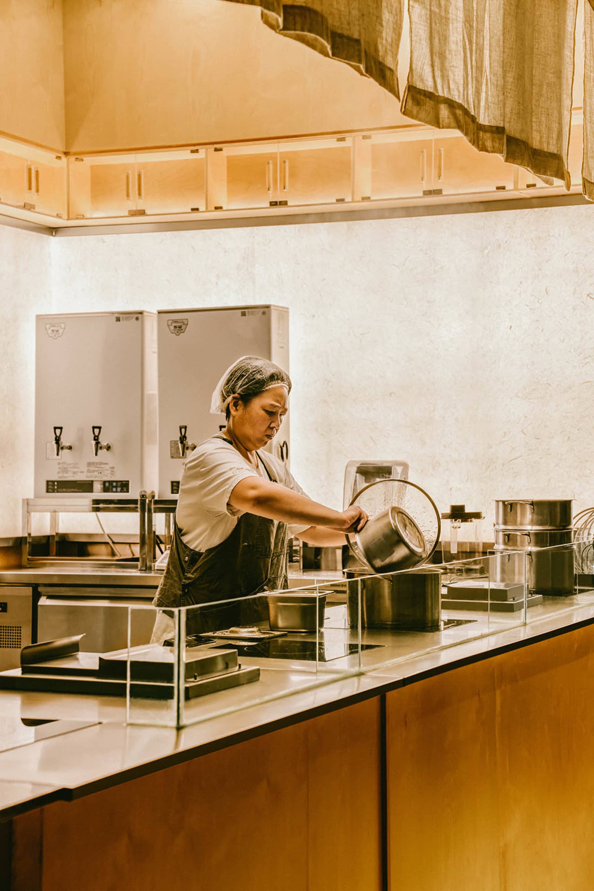 A woman in an industrial kitchen prepares food on a countertop, surrounded by bowls and utensils, in a professional environment with attention to detail