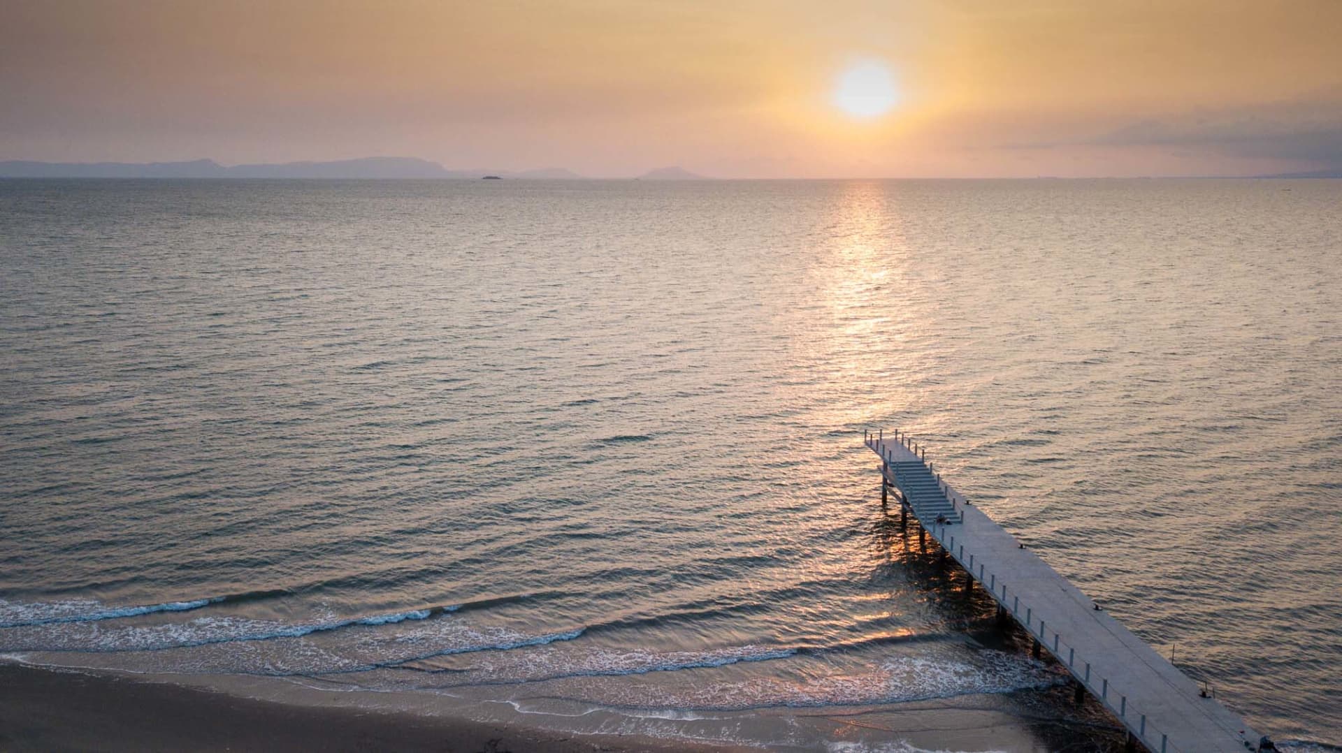 A wooden pier extends into the ocean at sunset, with a railing and a serene atmosphere created by the warm hues of the sky and the calm reflective water