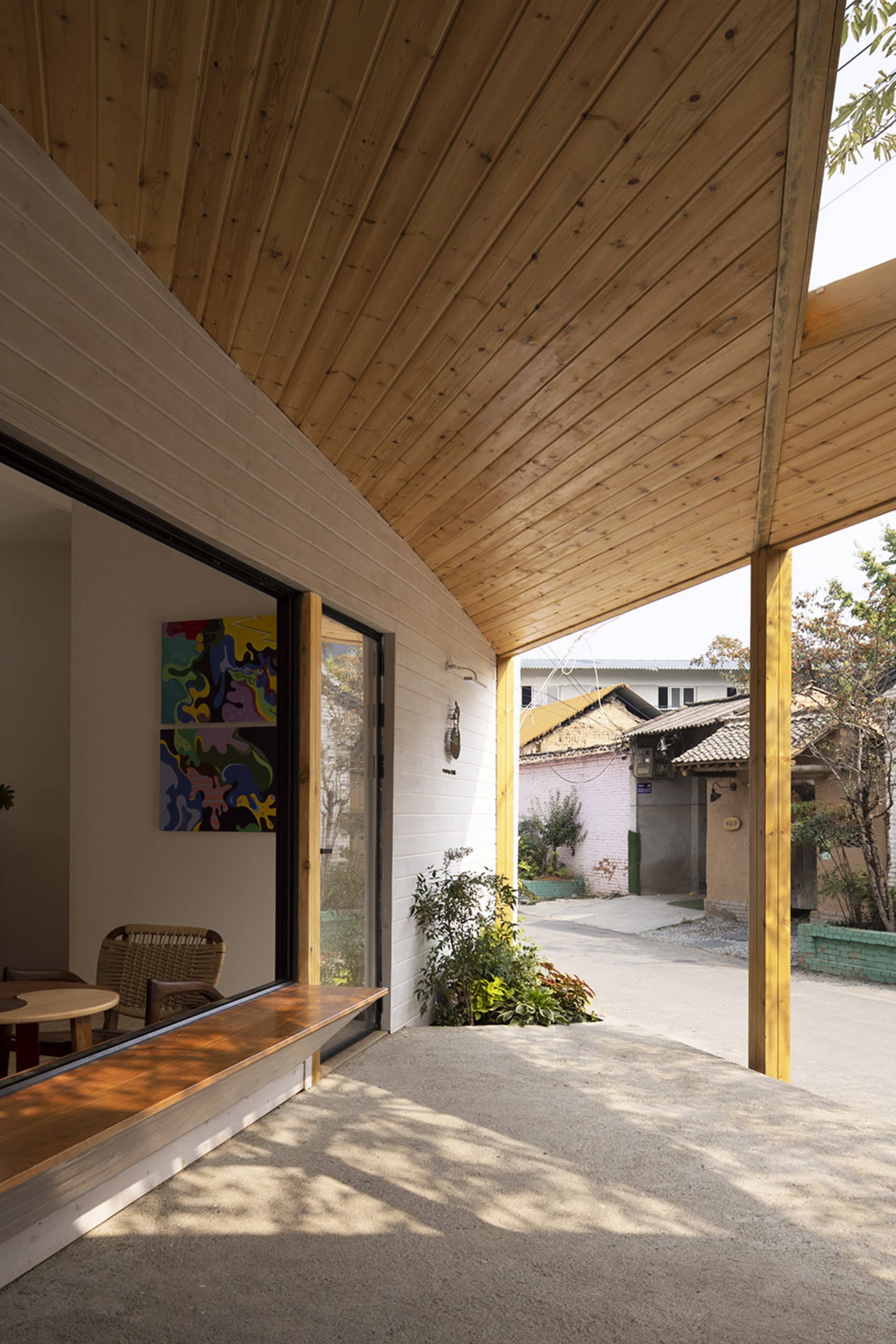 Modern house with wooden walls, a white roof, and a patio area with a bench and dining table, surrounded by chairs and a living room with a couch and potted plant