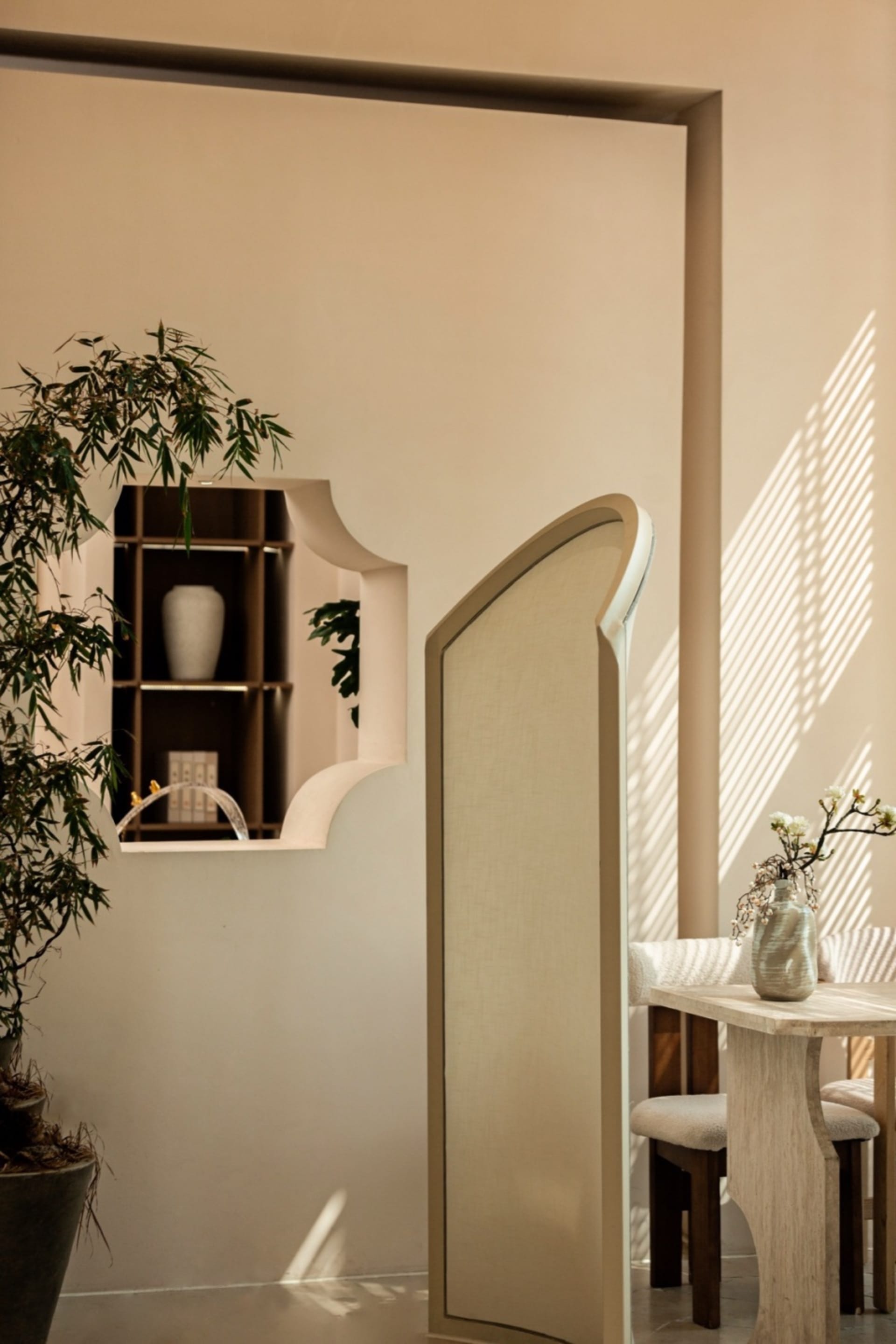 A bright and inviting interior space with a white wall, a large mirror, two potted plants, and a vase, lit by natural sunlight