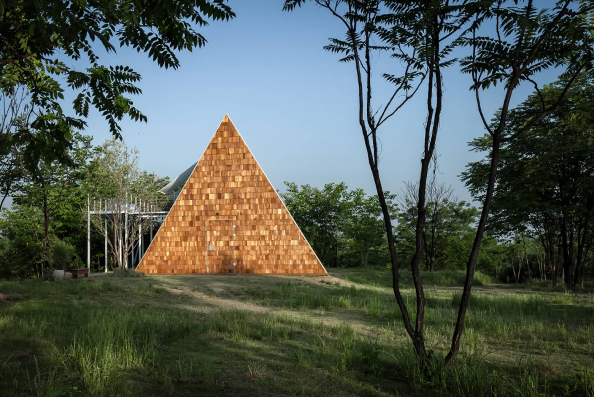 A unique wooden house with a triangular shape, surrounded by trees and grass in a serene natural setting