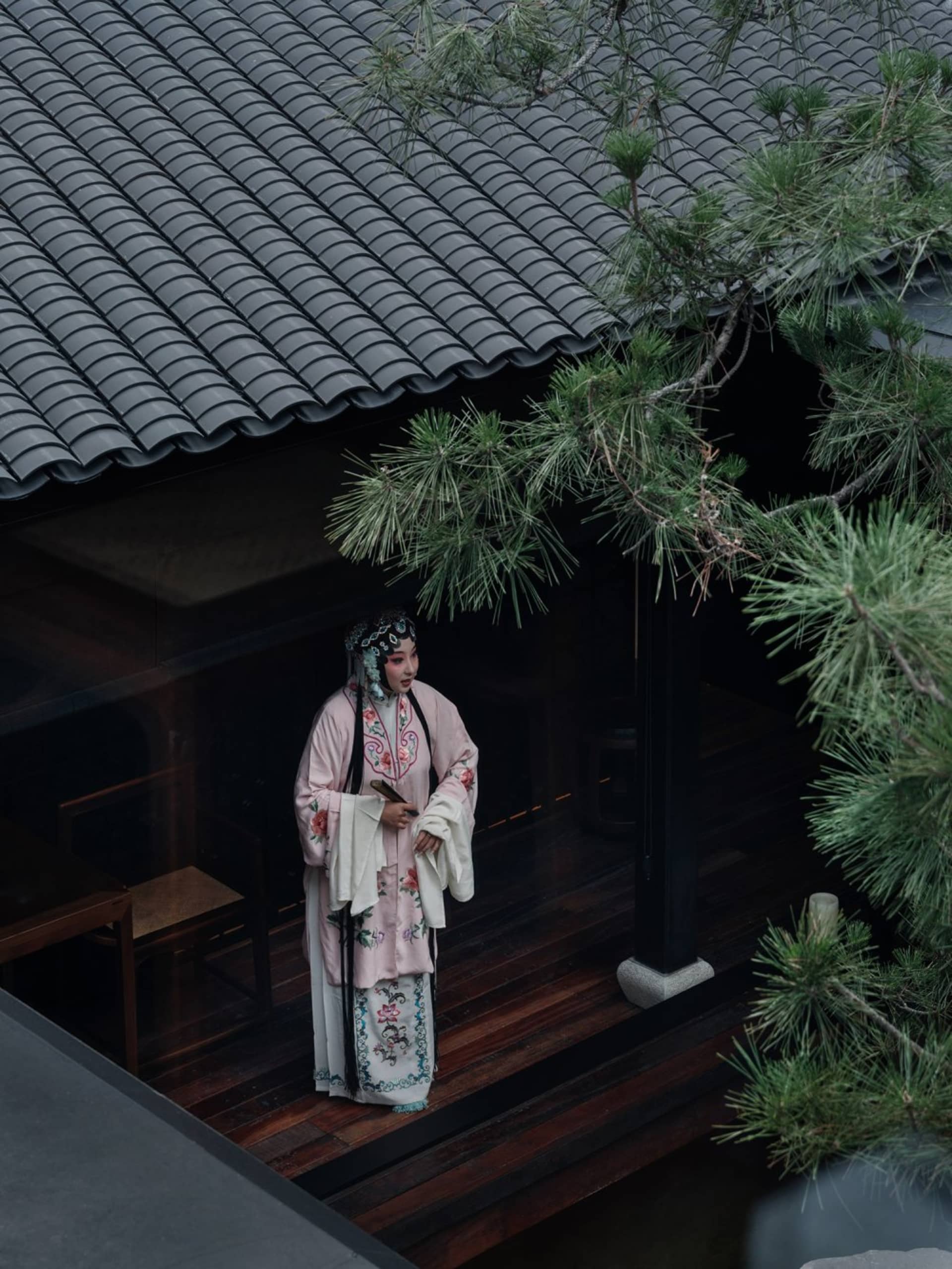 A woman stands in front of a traditional building with a wooden roof, surrounded by outdoor seating and suggesting cultural heritage