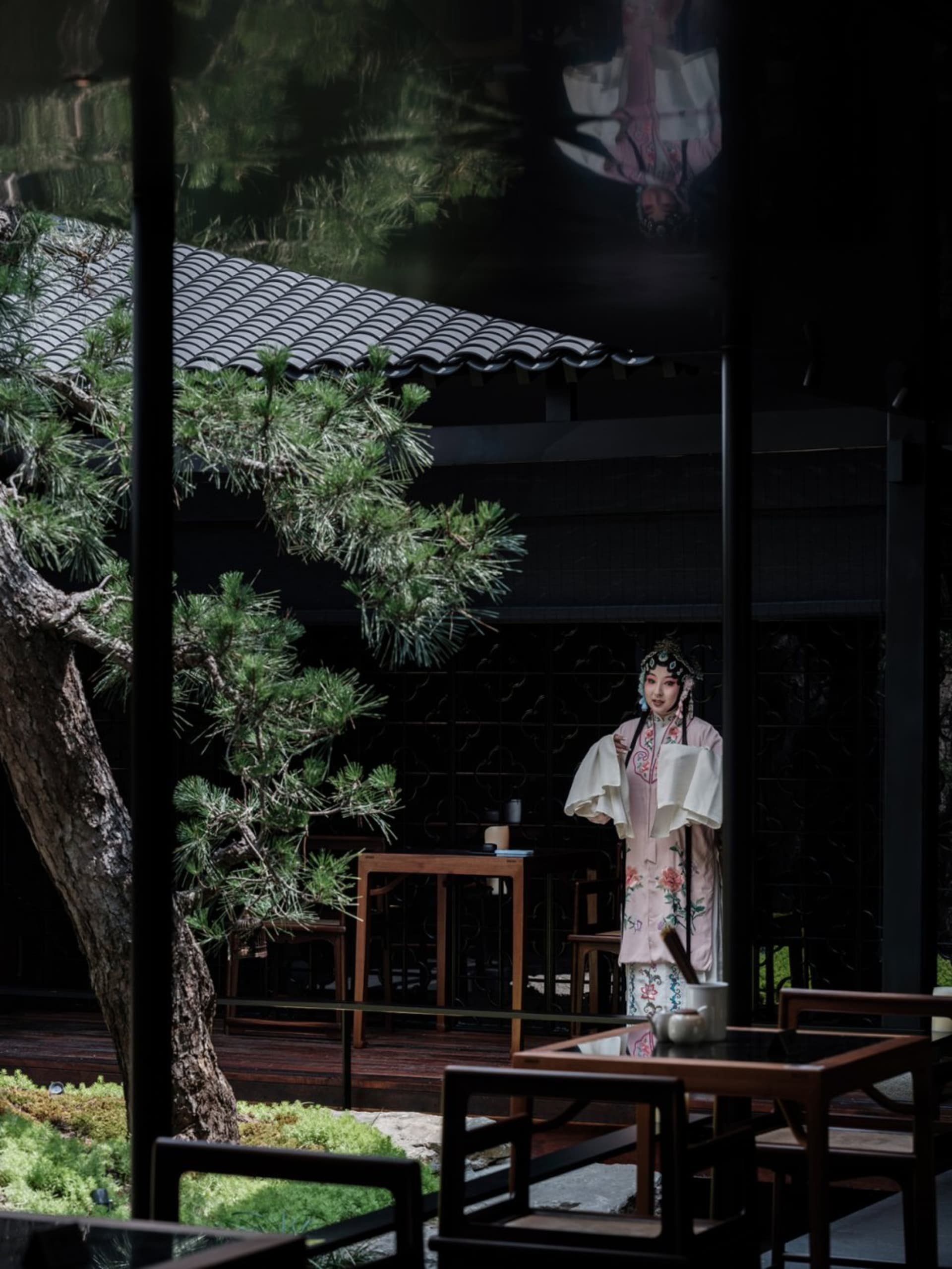A woman in traditional Chinese clothing stands in front of a table with a vase, surrounded by chairs in a restaurant setting, conveying cultural heritage and hospitality