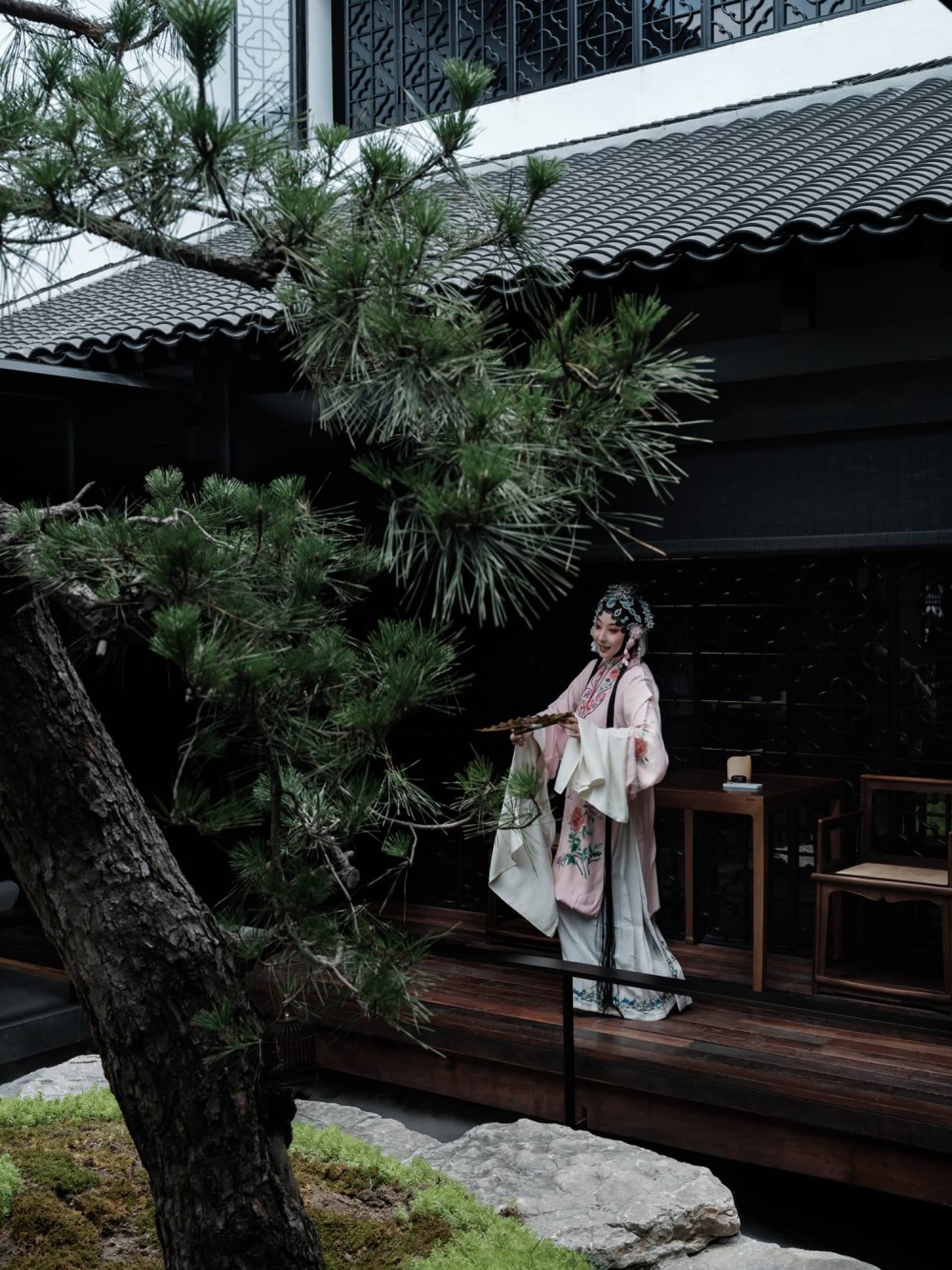 A woman in traditional attire stands in front of a building with a wooden table and chairs, surrounded by potted plants, conveying a sense of tradition and cultural significance