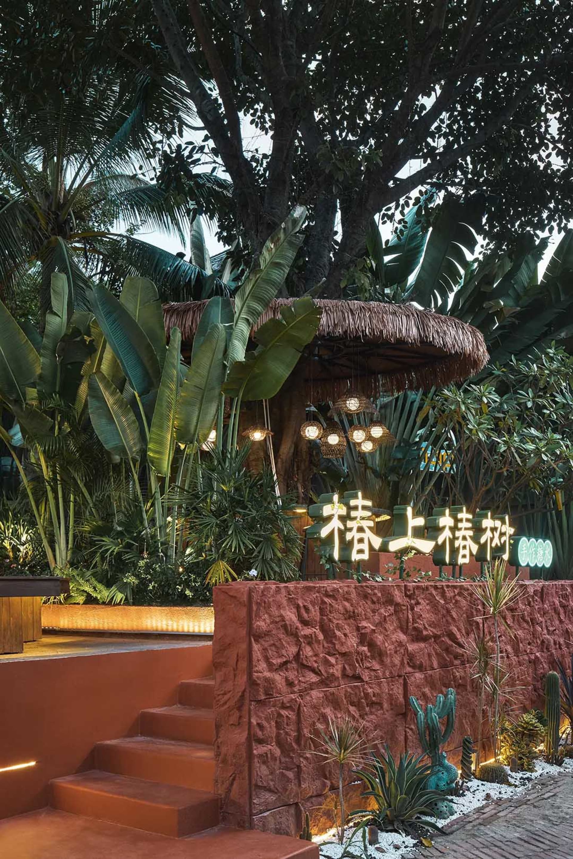 A serene outdoor patio area with a red brick wall, staircase, and small hut-like structure surrounded by palm trees and potted plants