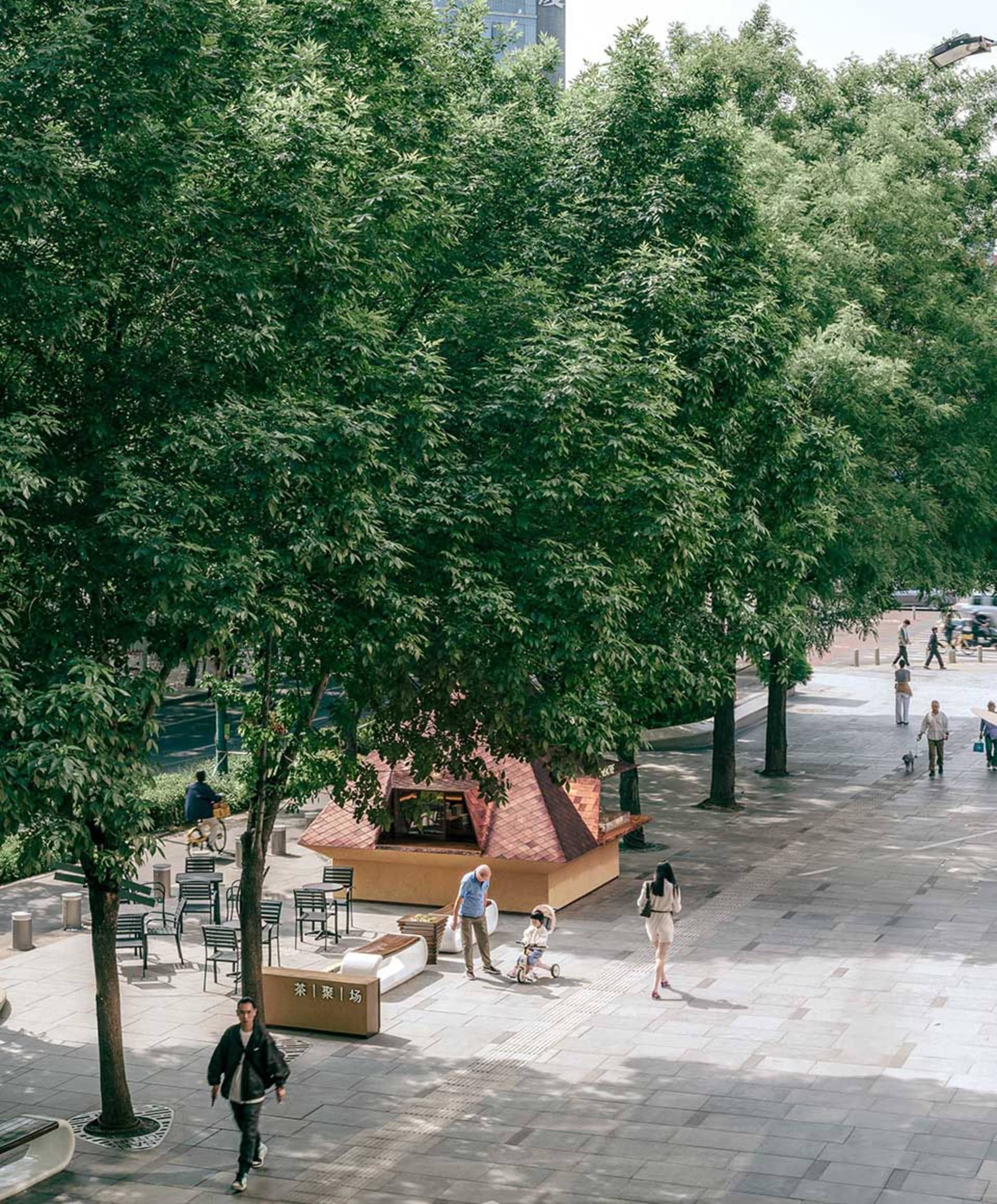 A bustling city street scene with a large tree, people walking, chairs, and umbrellas