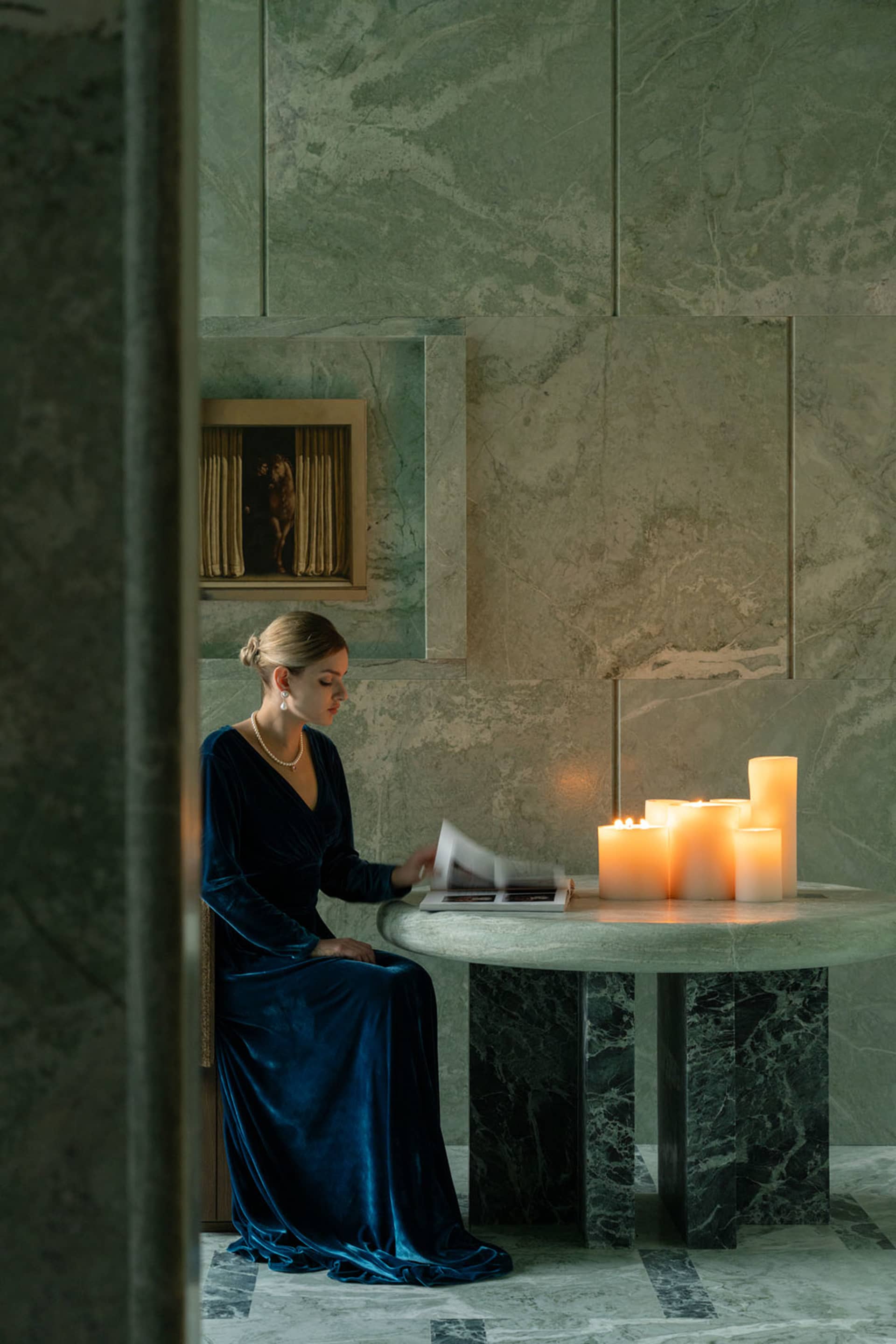 A woman sits at a table in a dimly lit room with a stone wall, surrounded by candles and a vase, with two chairs and a book