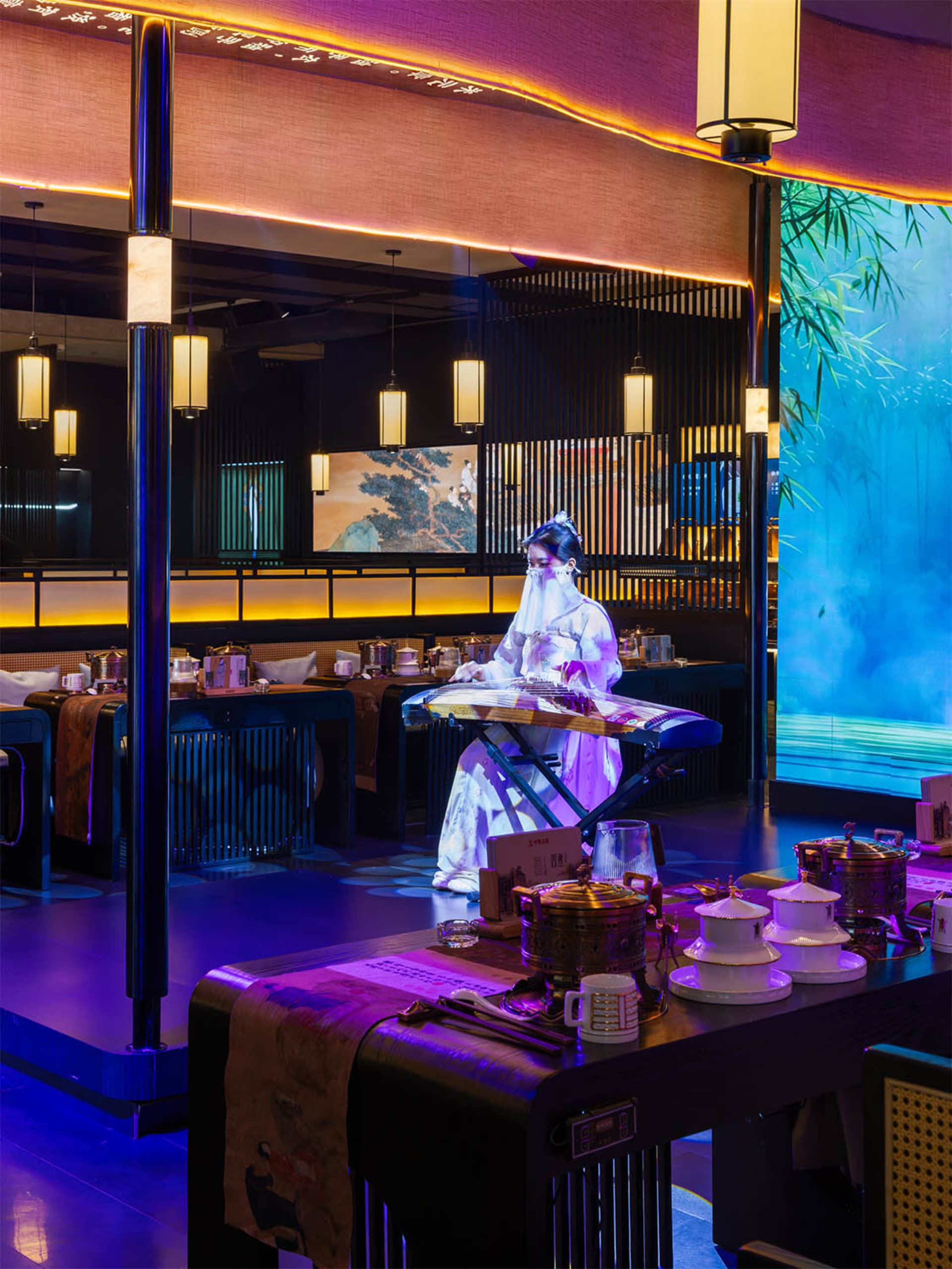 A woman enjoys her meal at a table in front of a large painting in a cozy Asian restaurant with multiple tables and chairs, cups on surfaces, and a relaxed atmosphere