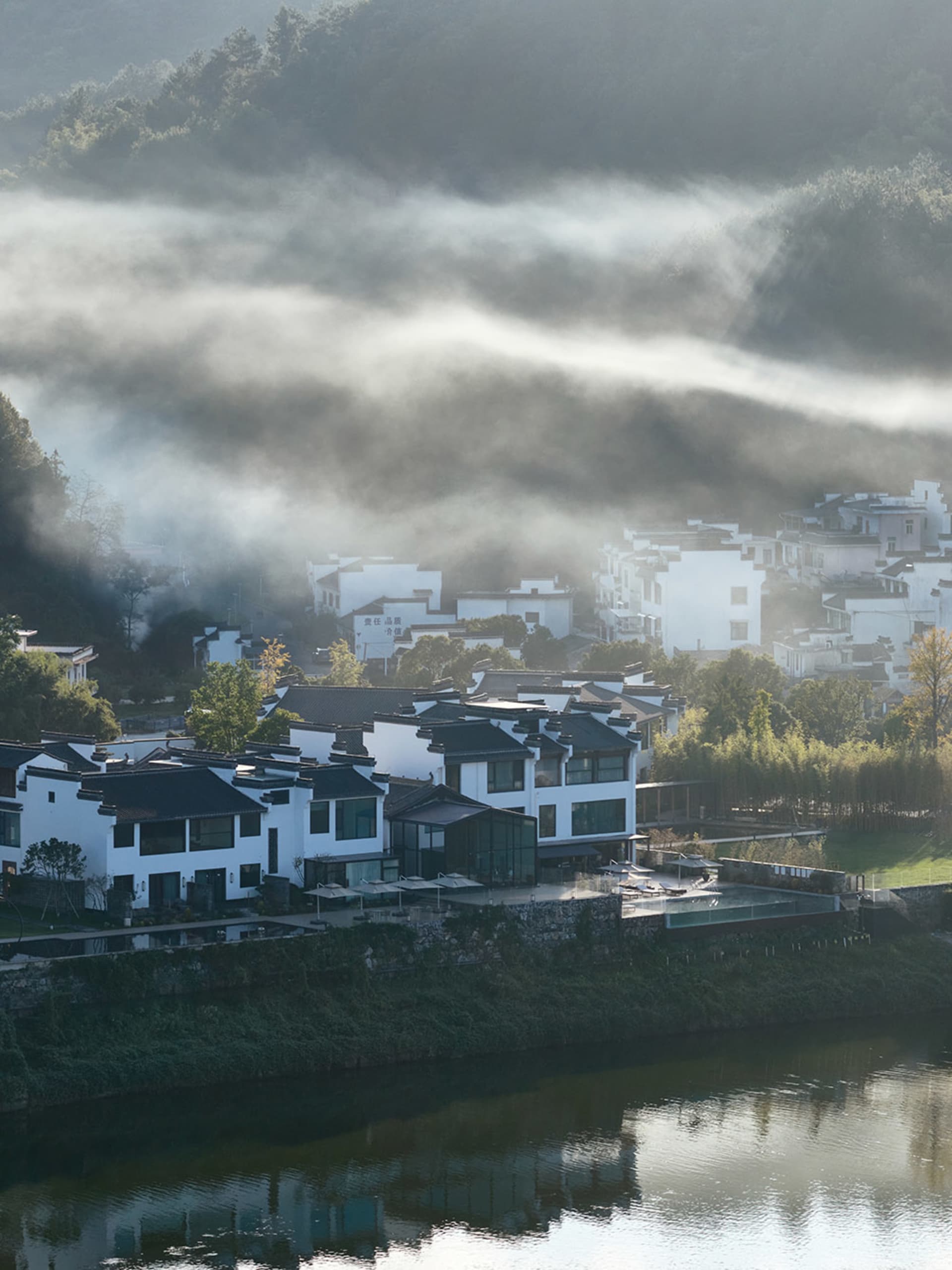 A serene Asian village with white houses on a hillside overlooking a river, with boats near the waterfront