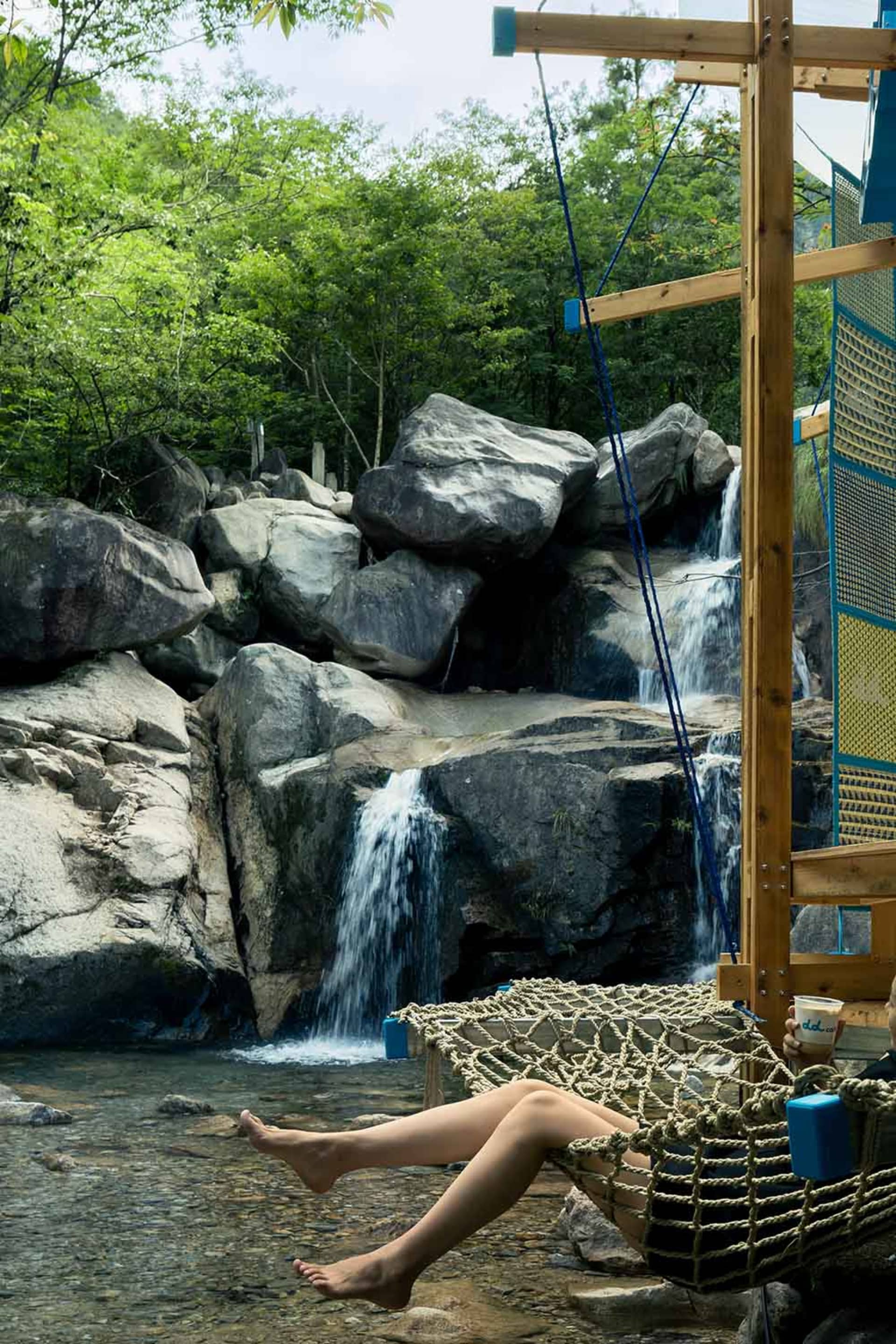 A serene outdoor scene featuring a waterfall surrounded by rocks and trees, with a wooden fence in the distance and two people present