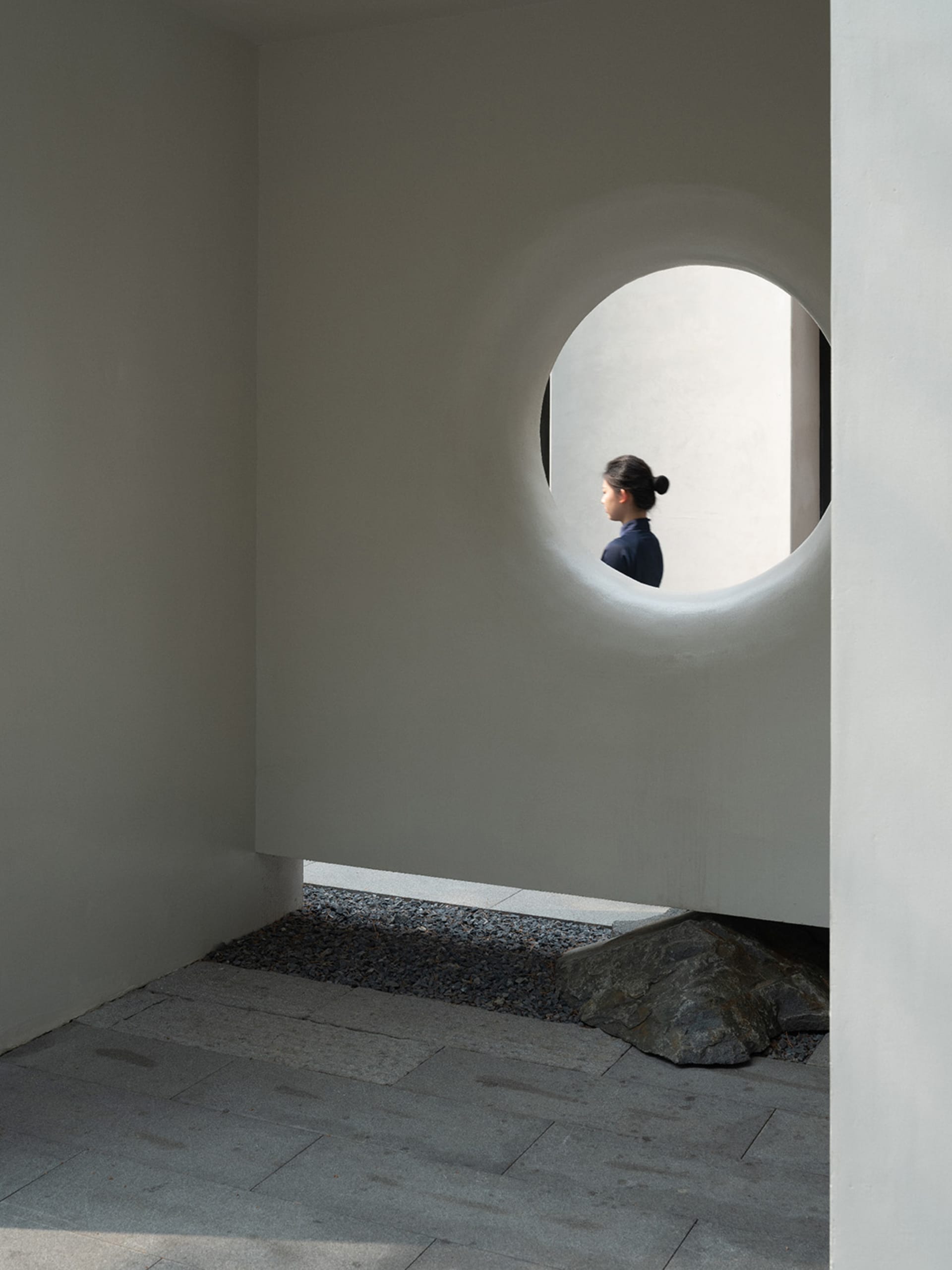 A woman stands in front of a concrete wall with a round window, surrounded by a calm and contemplative atmosphere