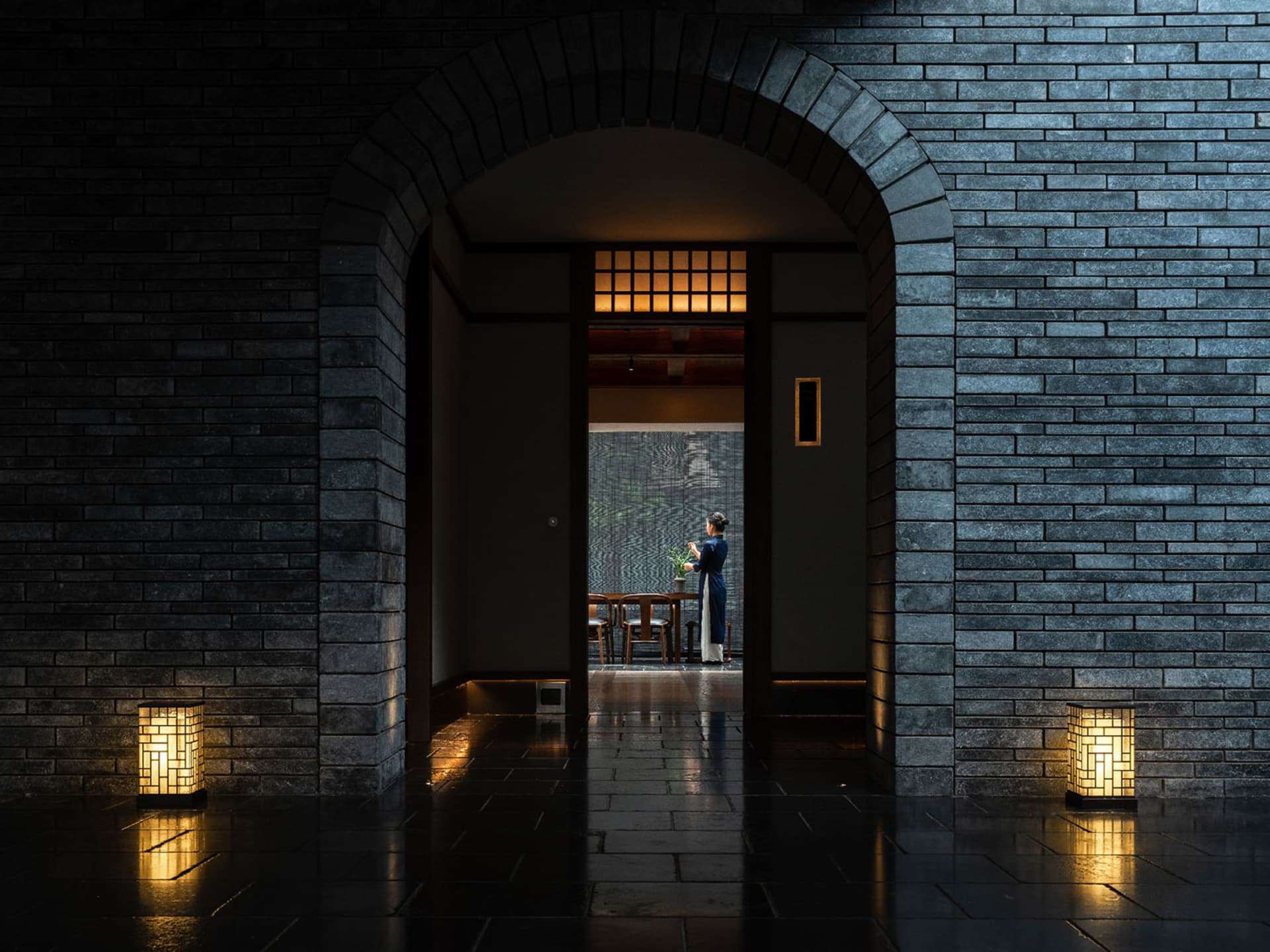 A woman stands in front of a doorway in a traditional Asian restaurant with stone walls, a brick archway, and cozy dining chairs