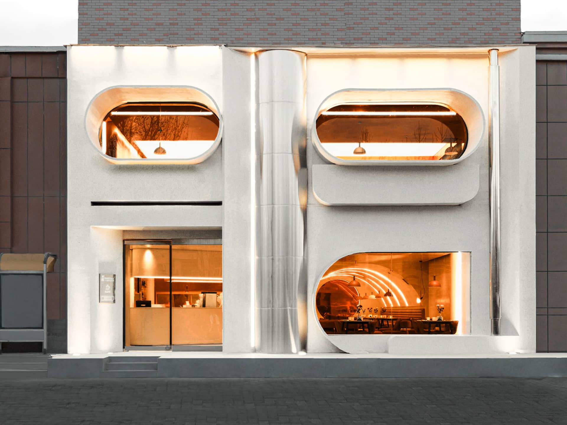 A modern building with a large lit window, white and grey elements, and outdoor seating areas, conveying elegance and sophistication