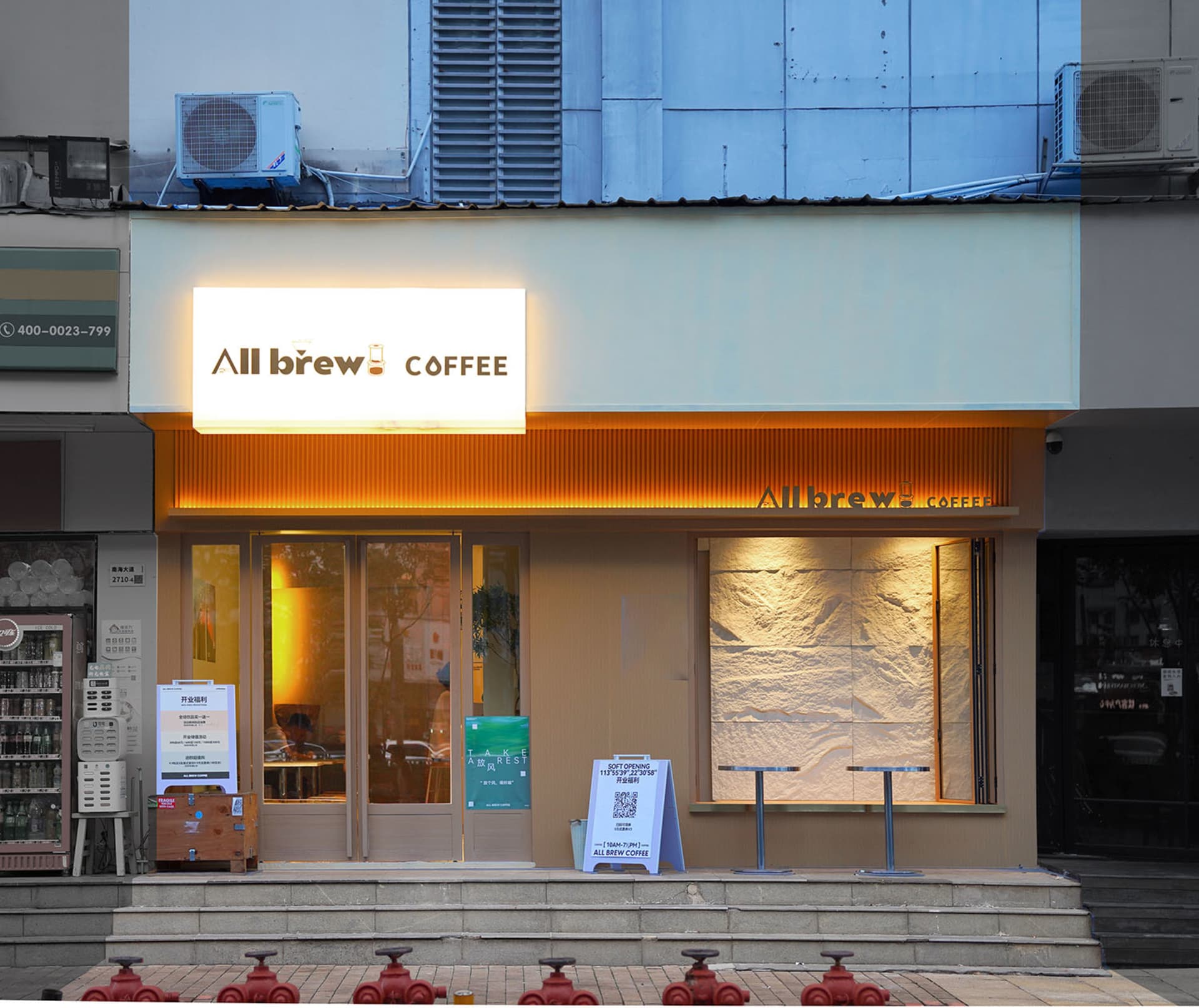 現代咖啡店入口，設有兩張餐桌、長椅與散置椅凳，周圍環繞白棕色牆面與盆栽