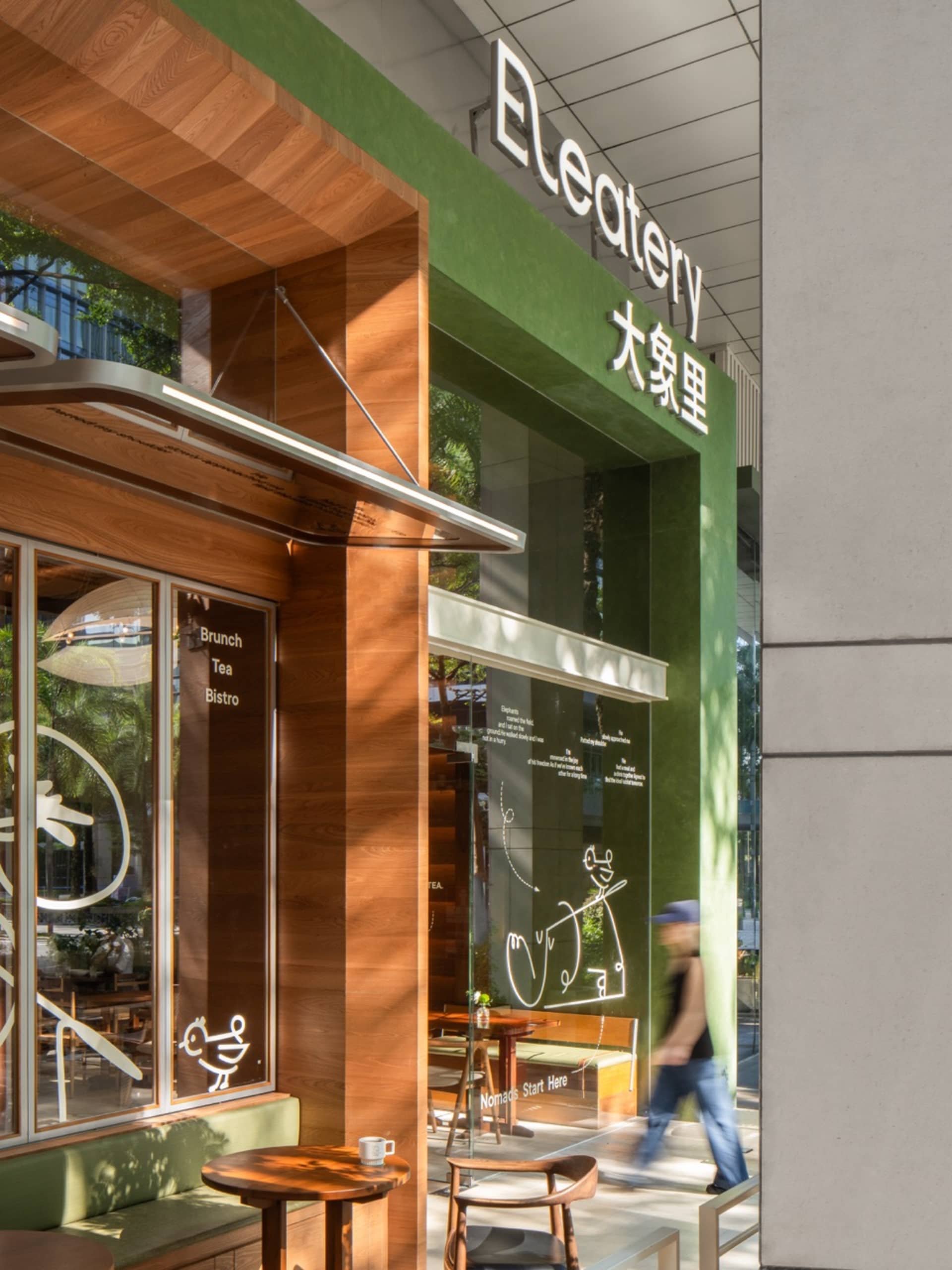 Cozy Asian restaurant with a wooden exterior, green accents, and large windows, featuring dining tables, chairs, umbrellas, and potted plants