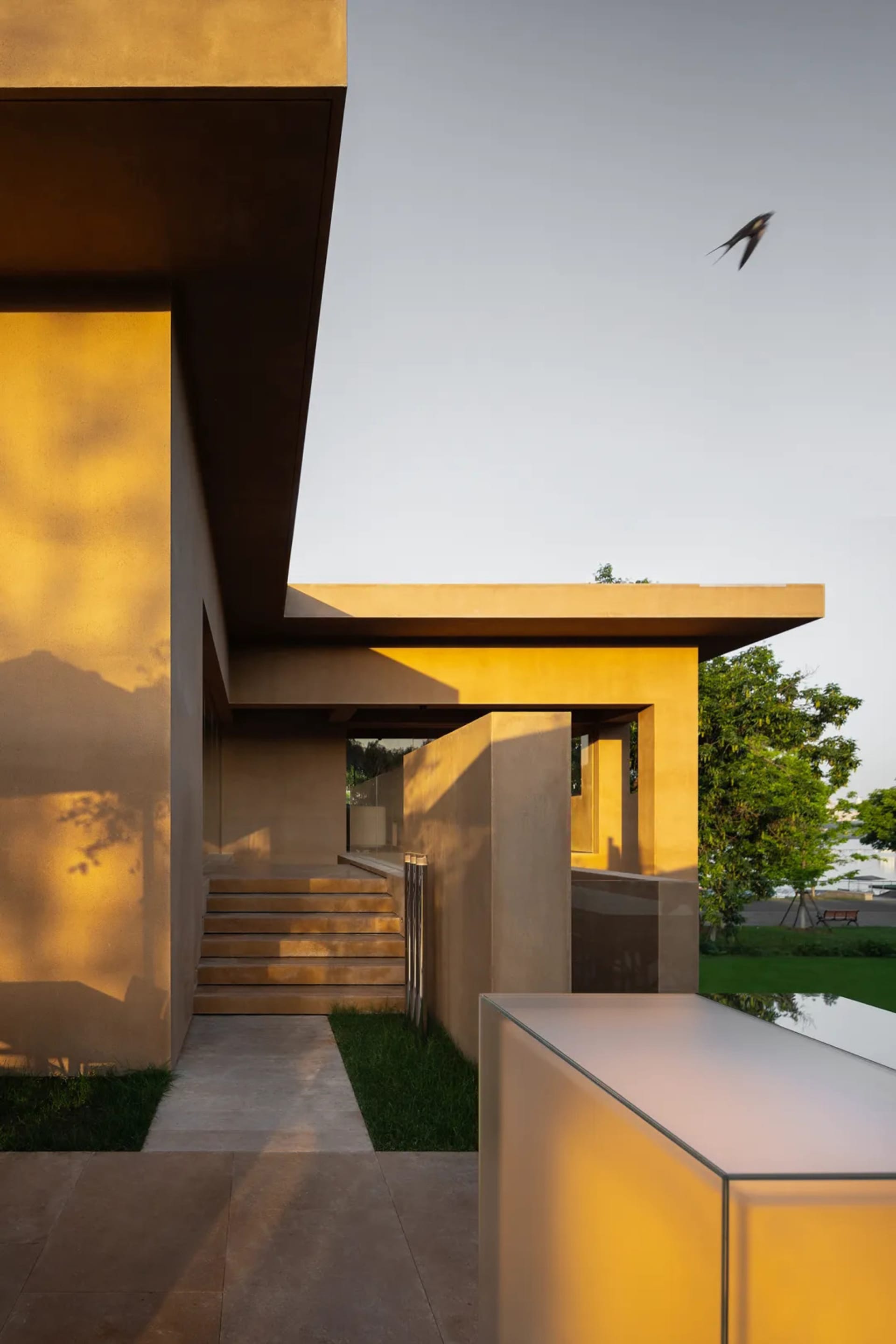 Modern house with a concrete exterior, glass windows, and a large patio area, featuring two staircases and a smaller building in the background, bathed in natural sunlight