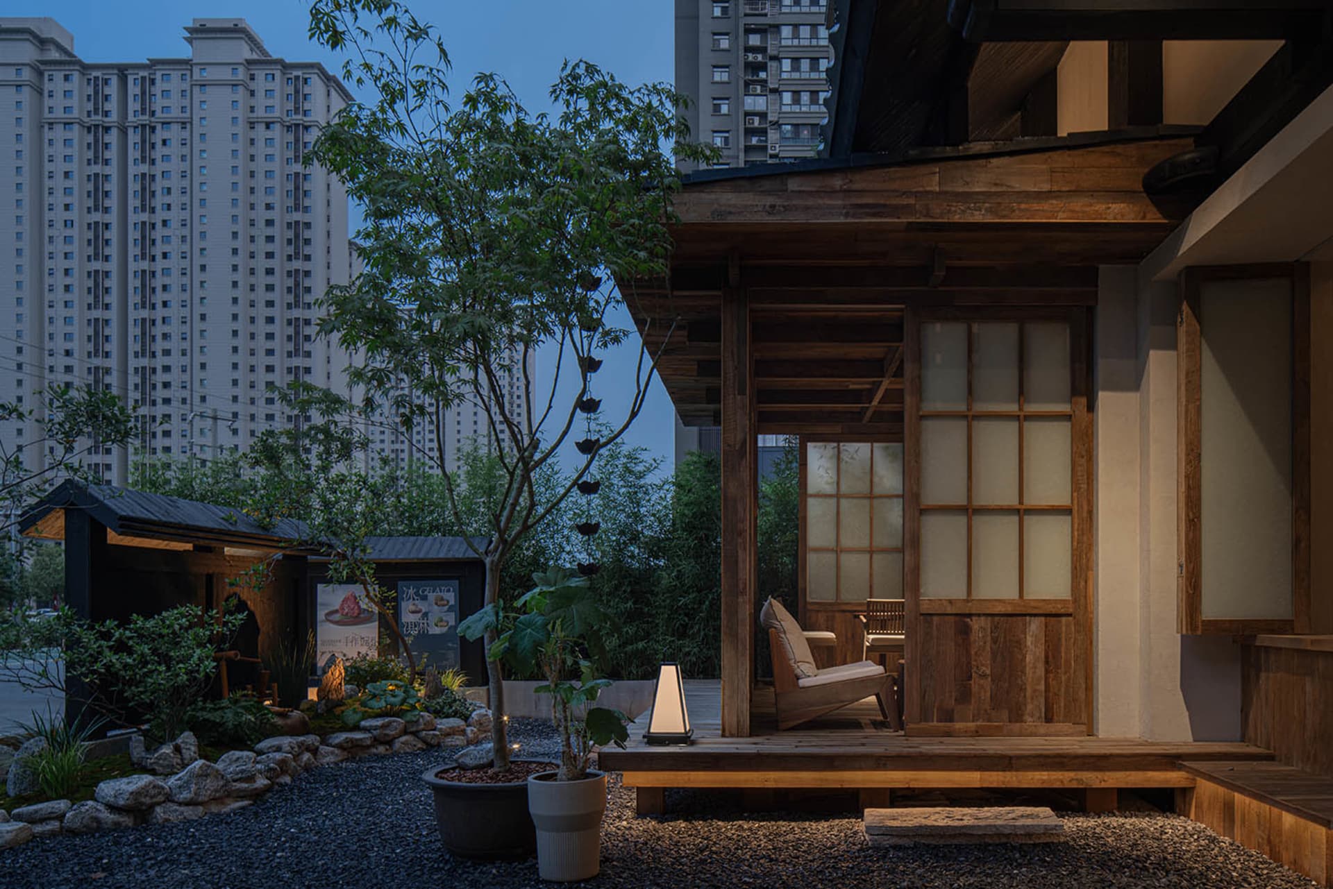 Asian-style house with wooden walls and a stone foundation, featuring a small sitting area with a couch and chair on a deck, surrounded by trees and a nighttime sky