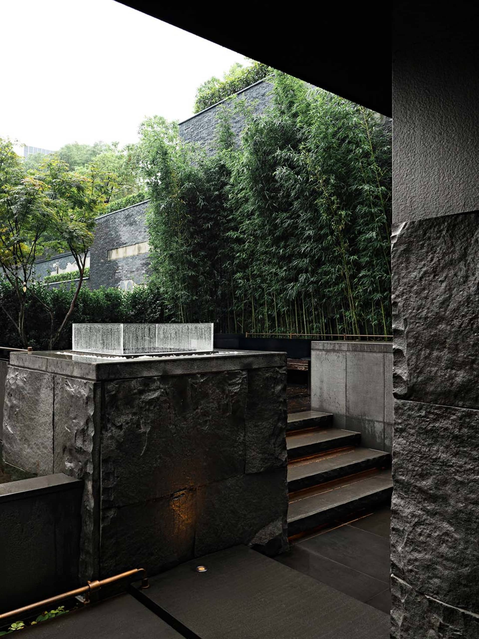 A modern living space with a stone wall featuring bamboo plants, a staircase, and potted plants throughout, blending indoor and outdoor elements