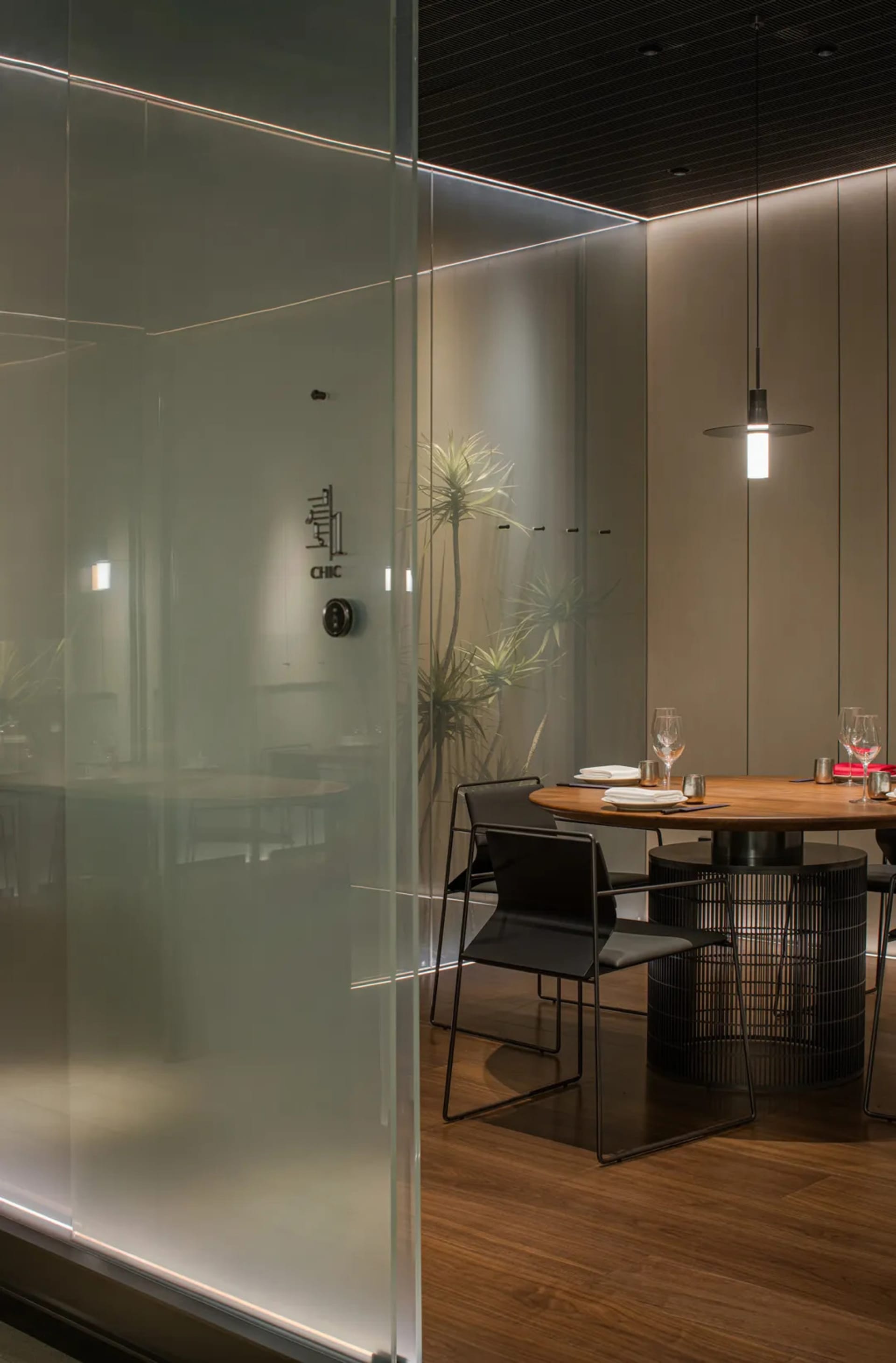 Modern dining space with a glass partition separating the kitchen from the dining area, featuring two chairs and a table set for a meal, with a smaller dining table in the background