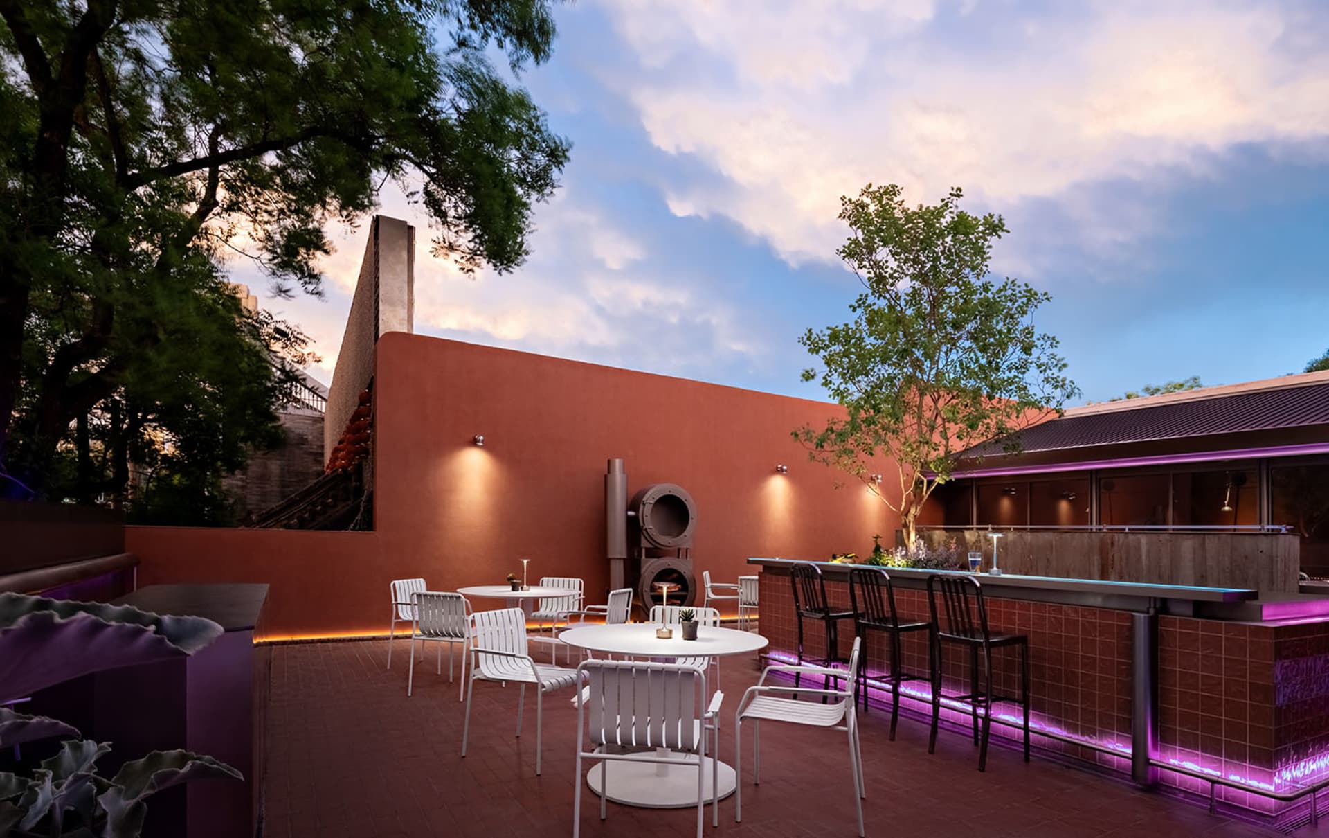 A cozy outdoor patio area with white chairs and tables around a central fire pit, set on a brick surface, surrounded by potted plants