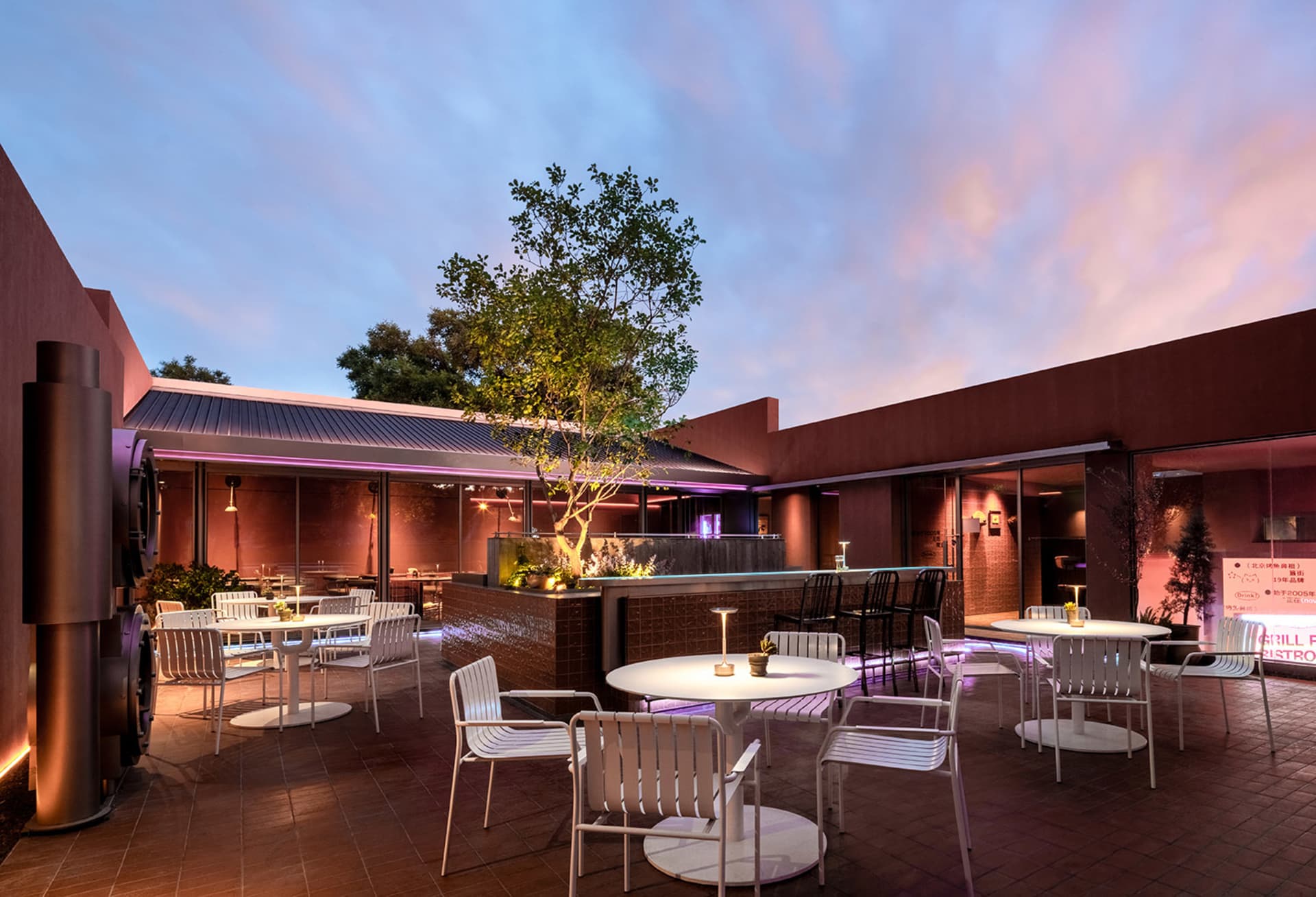 A restaurant patio area with a dining table and chairs, surrounded by potted plants and a tree, creating a relaxed and inviting atmosphere