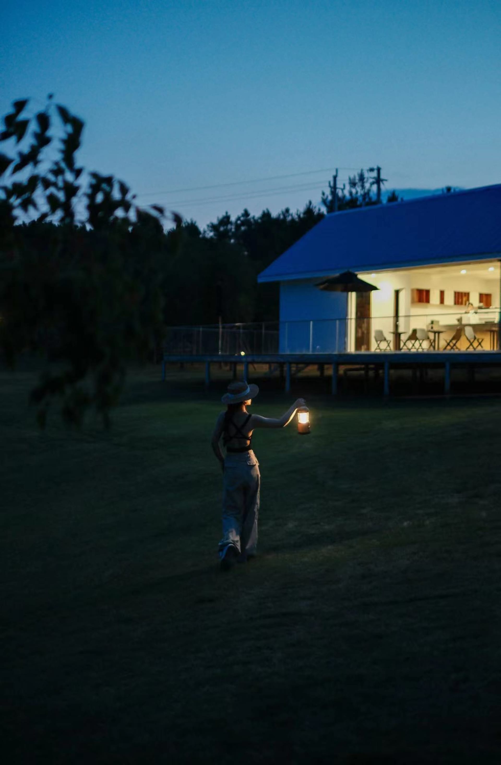 A woman walks across a grassy field at night, holding a lantern, surrounded by chairs and a dining table in a peaceful outdoor setting