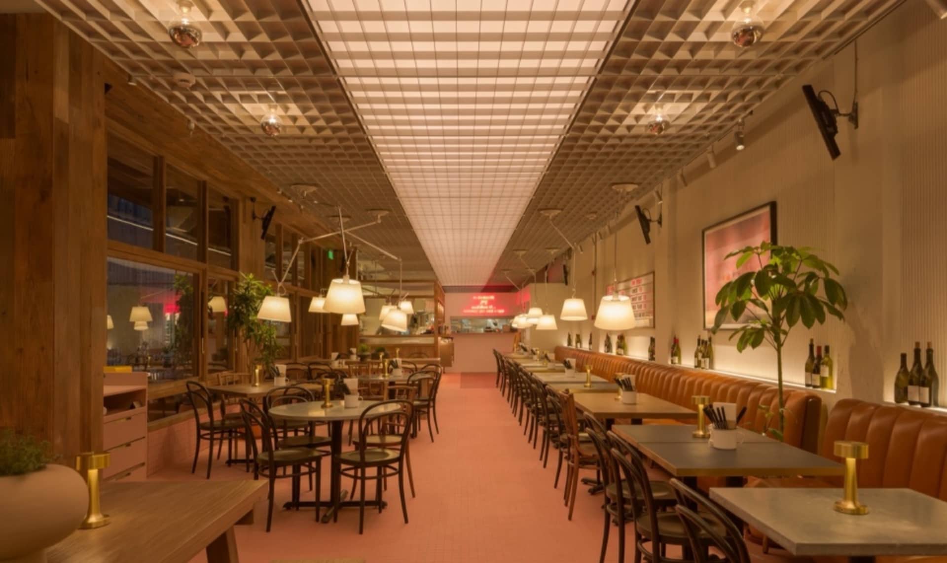 A modern restaurant interior with a long dining table, pink carpet, and potted plants, featuring wine glasses and a well-lit atmosphere