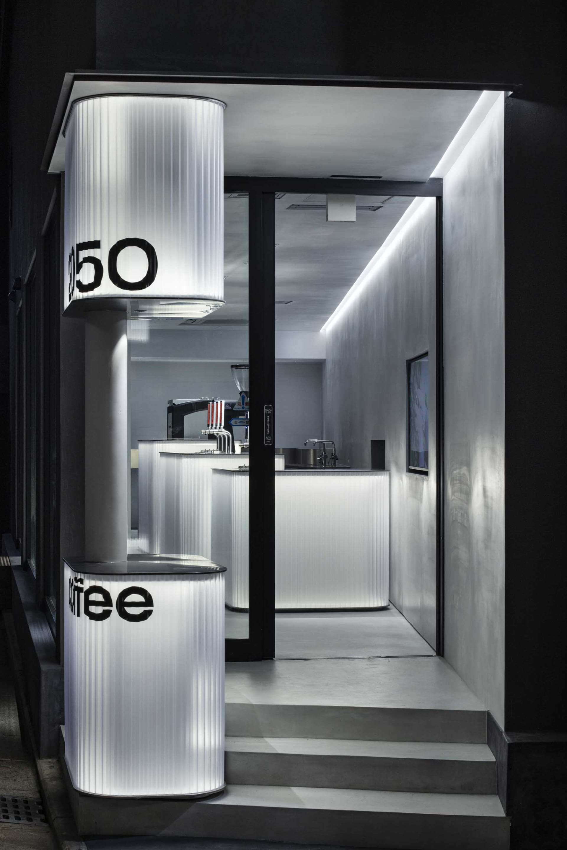 A modern industrial cafe with concrete walls, a counter area, and a black door leading outside, featuring a coffee machine and cups on the countertop
